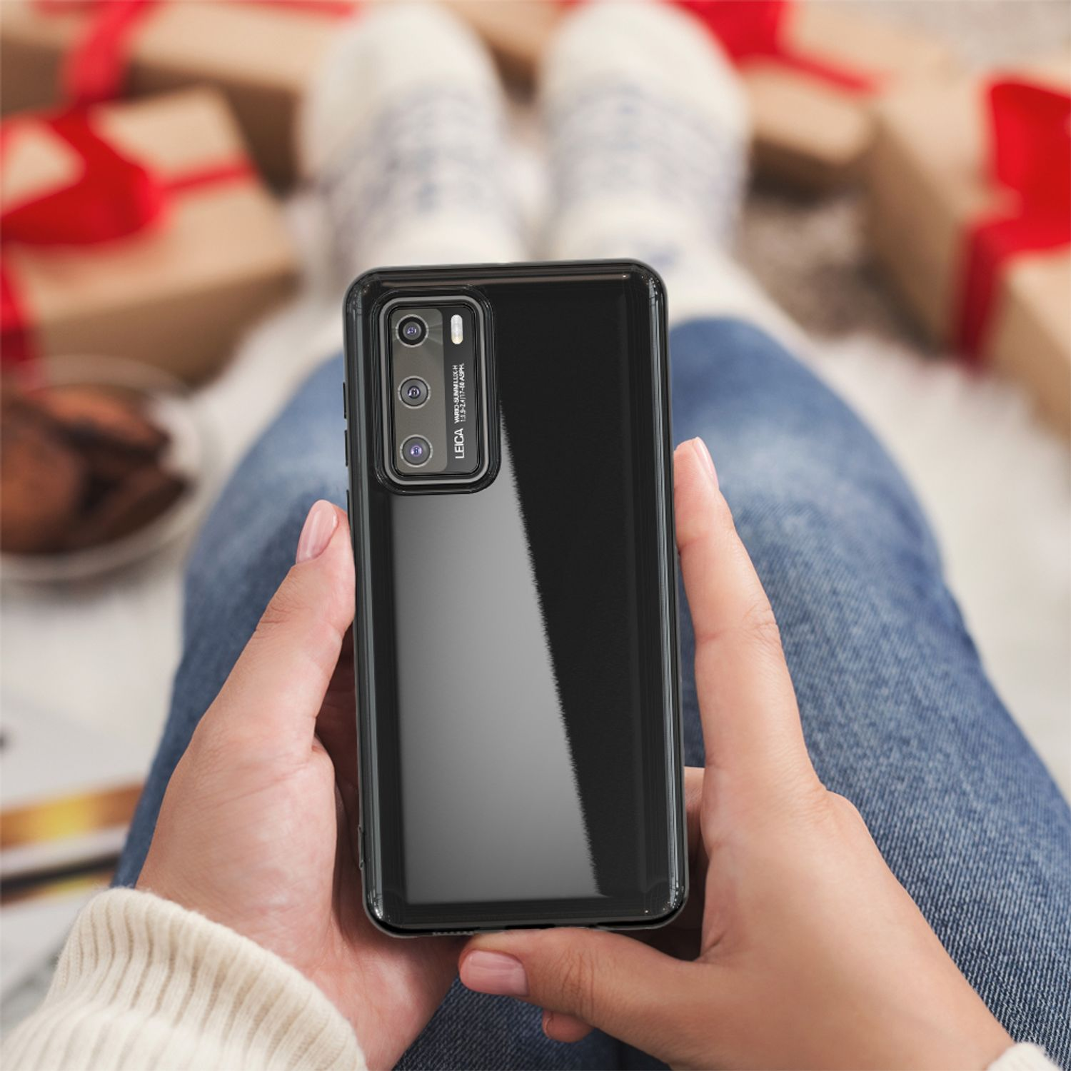 Hände halten ein schwarzes Telefon in einer Hülle. Eine Person sitzt mit hochgezogenen Beinen, umgeben von Geschenken.