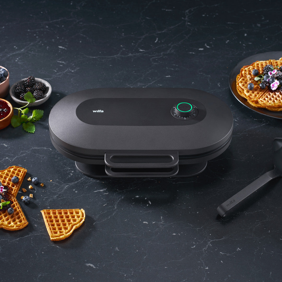 Macchina per waffle nera con waffle. Waffle, bacche e sciroppo su una superficie scura.