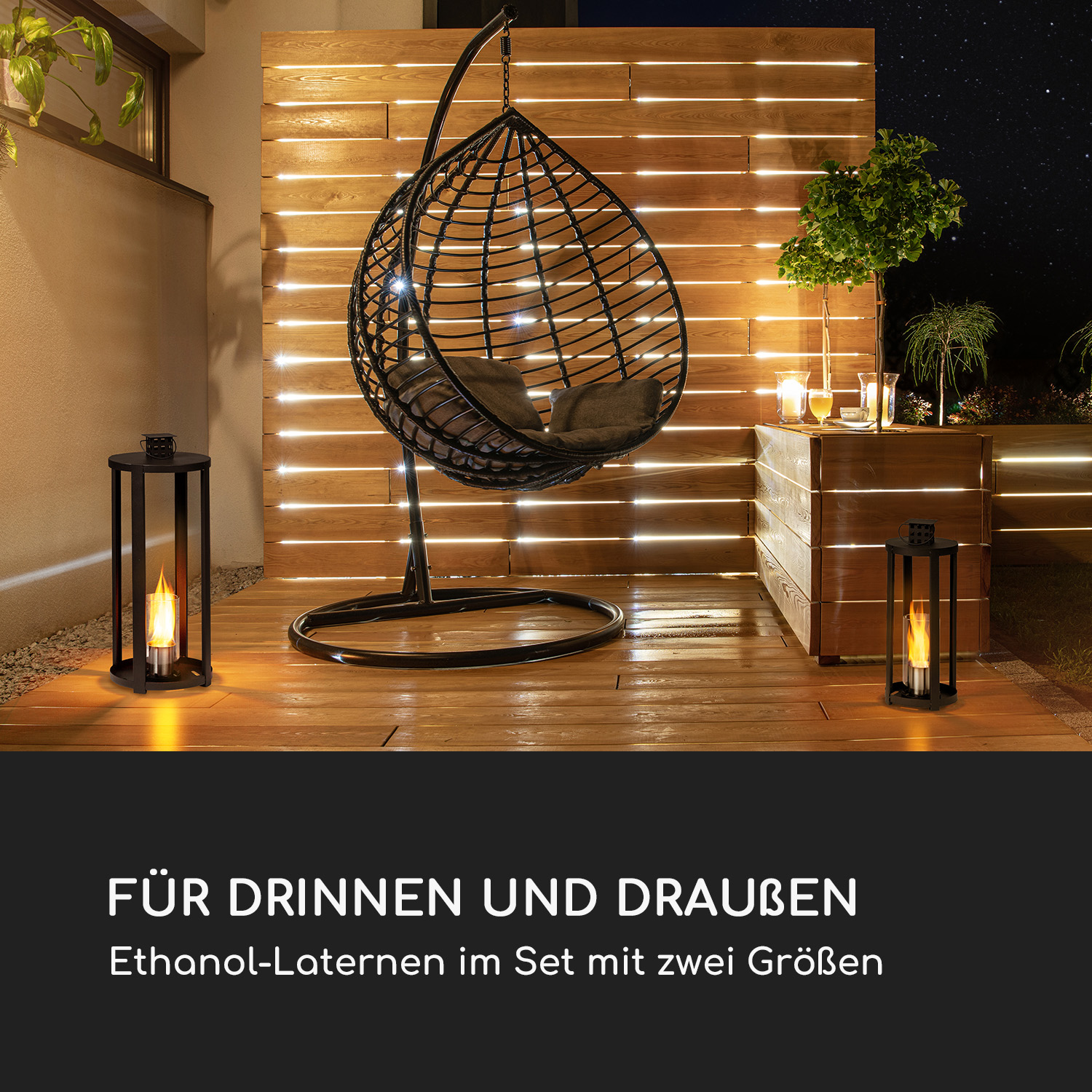 Eine Terrasse bei Nacht mit einem Hängesessel, zwei Laternen mit Flammen und einem Holzzaun mit Lichtern.