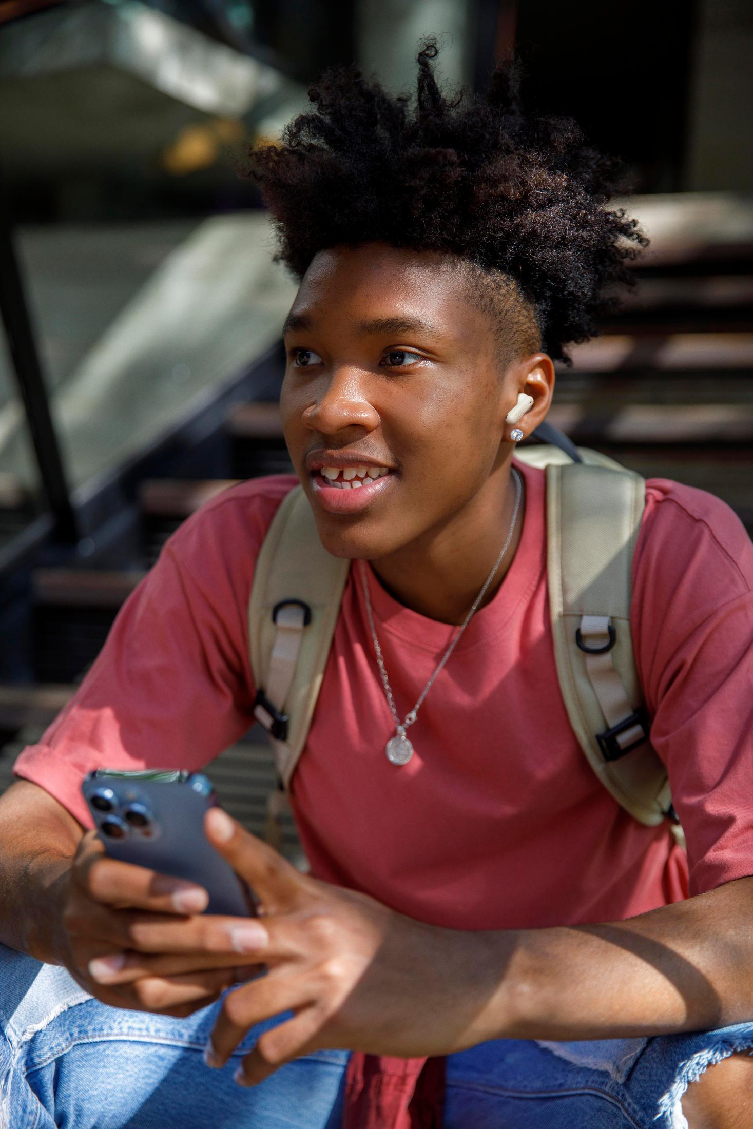 Młody czarnoskóry mężczyzna ze słuchawkami, trzymający telefon, ubrany w różową koszulę i plecak.