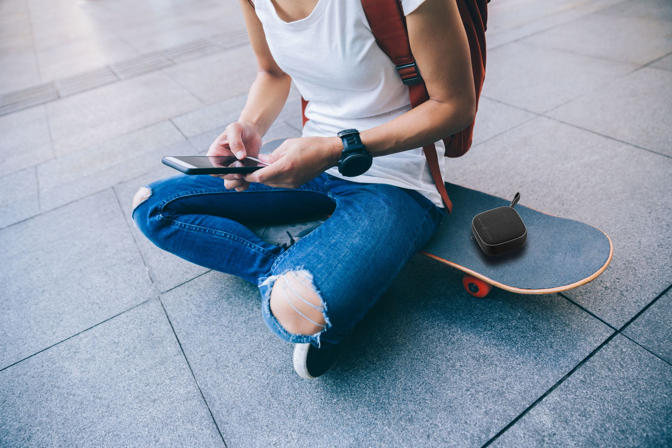 Osoba siedząca na deskorolce, używająca smartfona, z małym głośnikiem obok.
