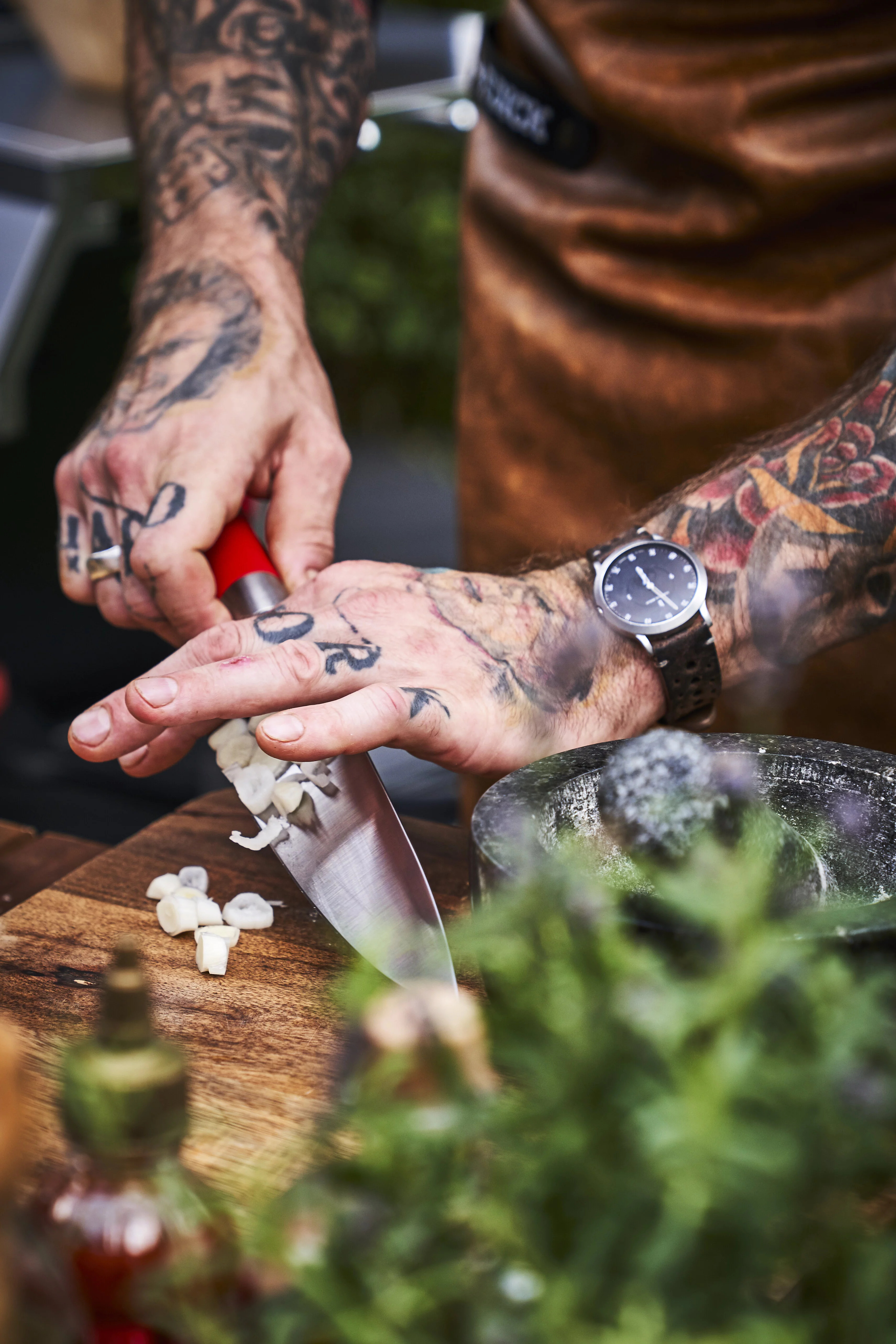 Ein tätowierter Koch hackt Knoblauch. Das Messer ist rot, das Schneidebrett aus Holz.