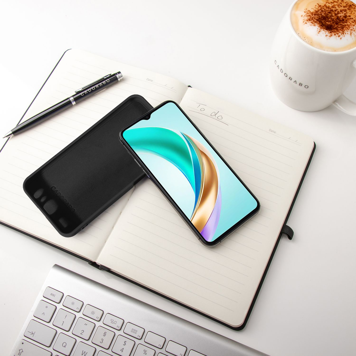 Notizbuch mit Telefon, Stift und Kaffeetasse auf einem weißen Schreibtisch. Telefon und Stift haben Logos.