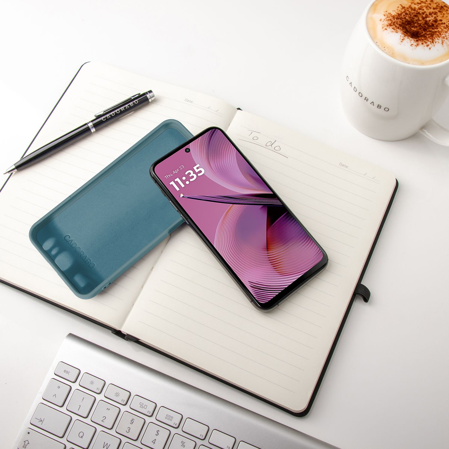 Smartphone auf Notizbuch mit Hülle, Stift und Kaffeetasse, auf weißem Schreibtisch.