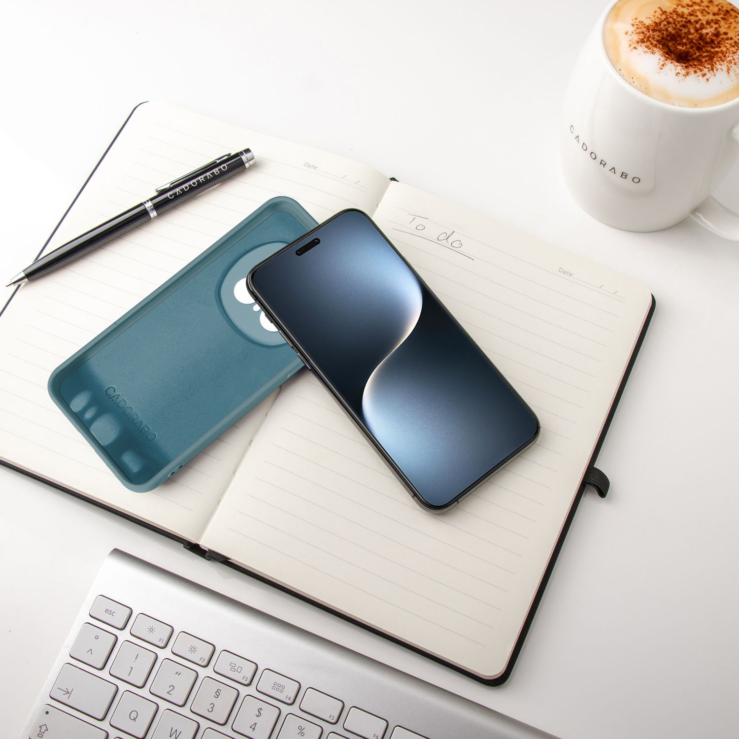 Telefon, türkisfarbene Hülle, Notizbuch, Stift und Kaffeetasse auf weißem Schreibtisch, Draufsicht.