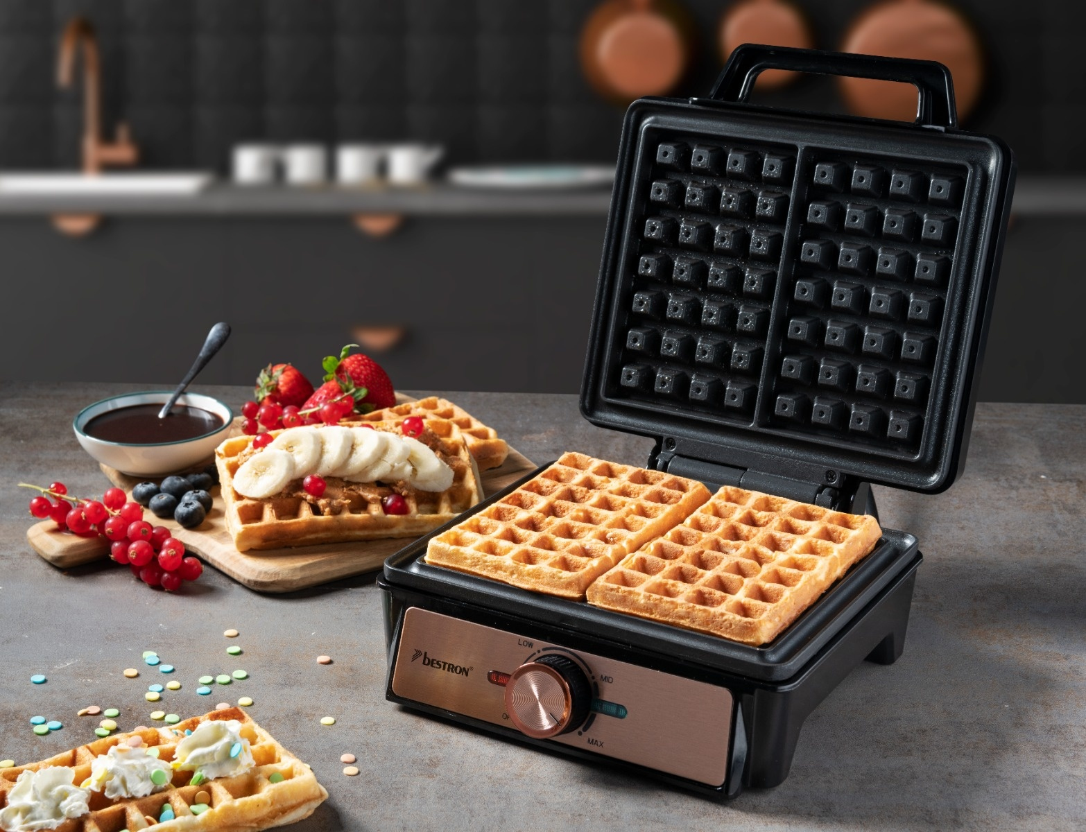 Una macchina per waffle Bestron con due waffle. Waffle su un tagliere con frutta. Salsa al cioccolato.