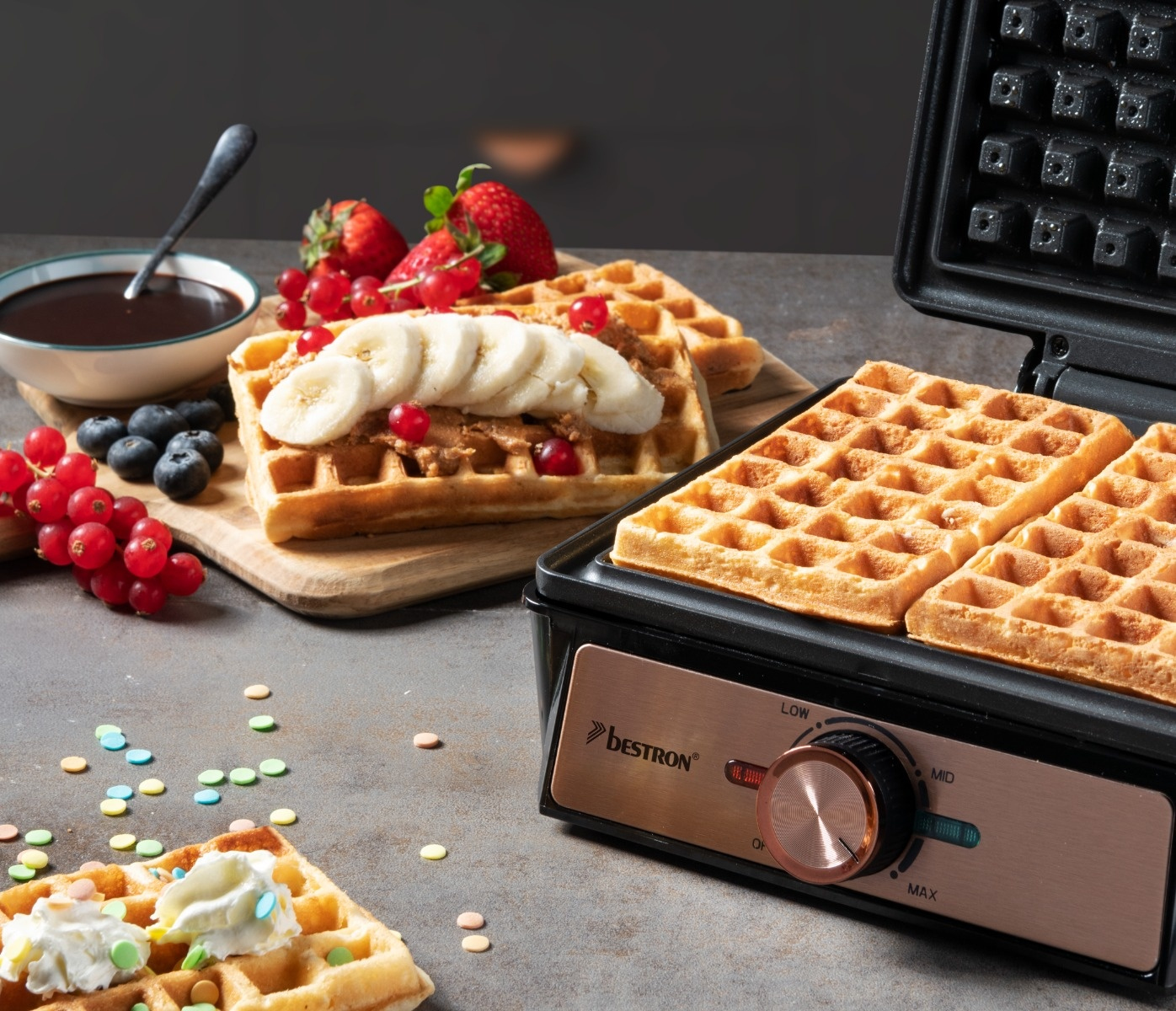 Macchina per waffle Bestron con waffle, frutta e salsa al cioccolato. Waffle su un tagliere. Zuccherini.