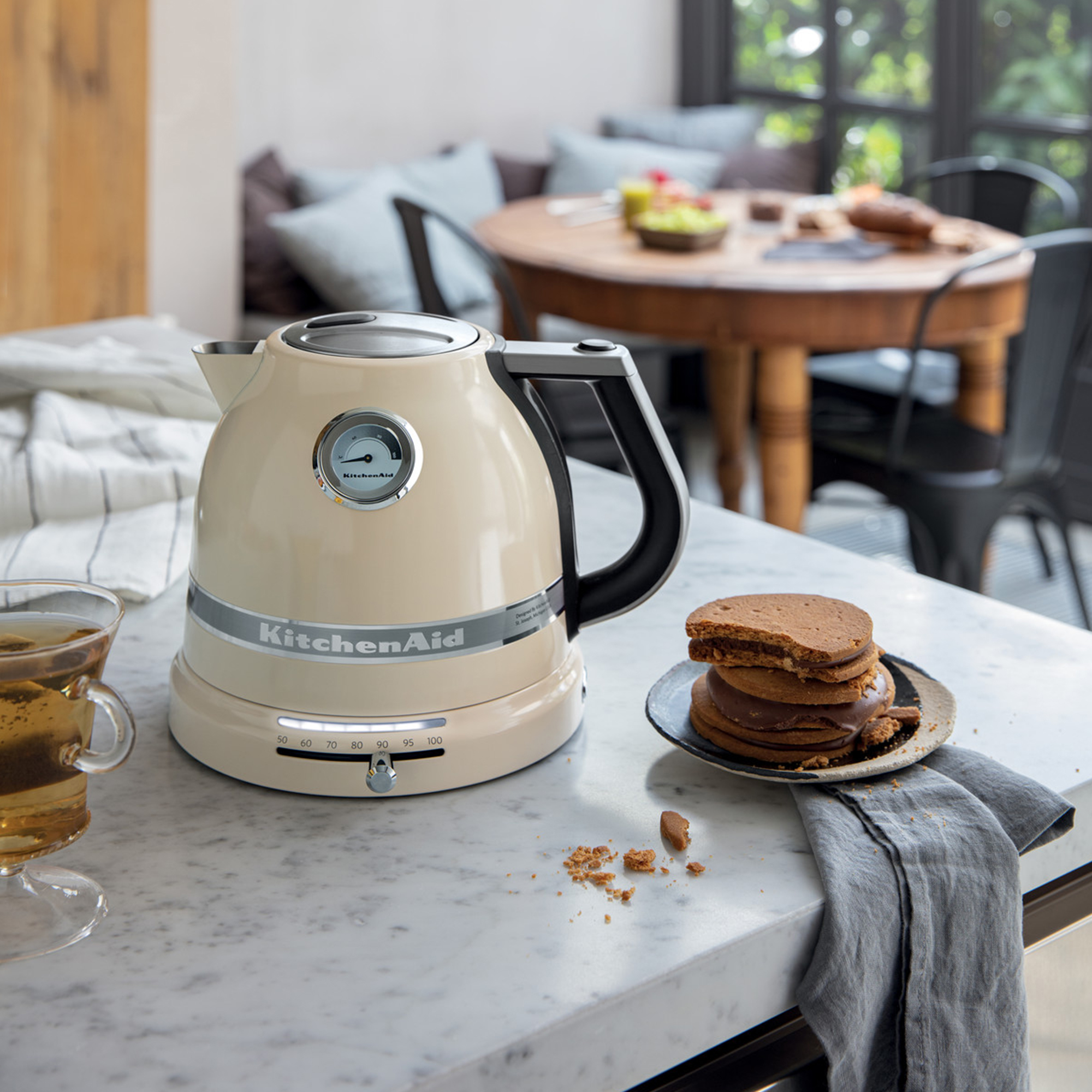 Cremefarbene KitchenAid-Kanne, Teetasse und Kekse auf der Theke, mit verschwommenem Hintergrund eines Essbereichs.