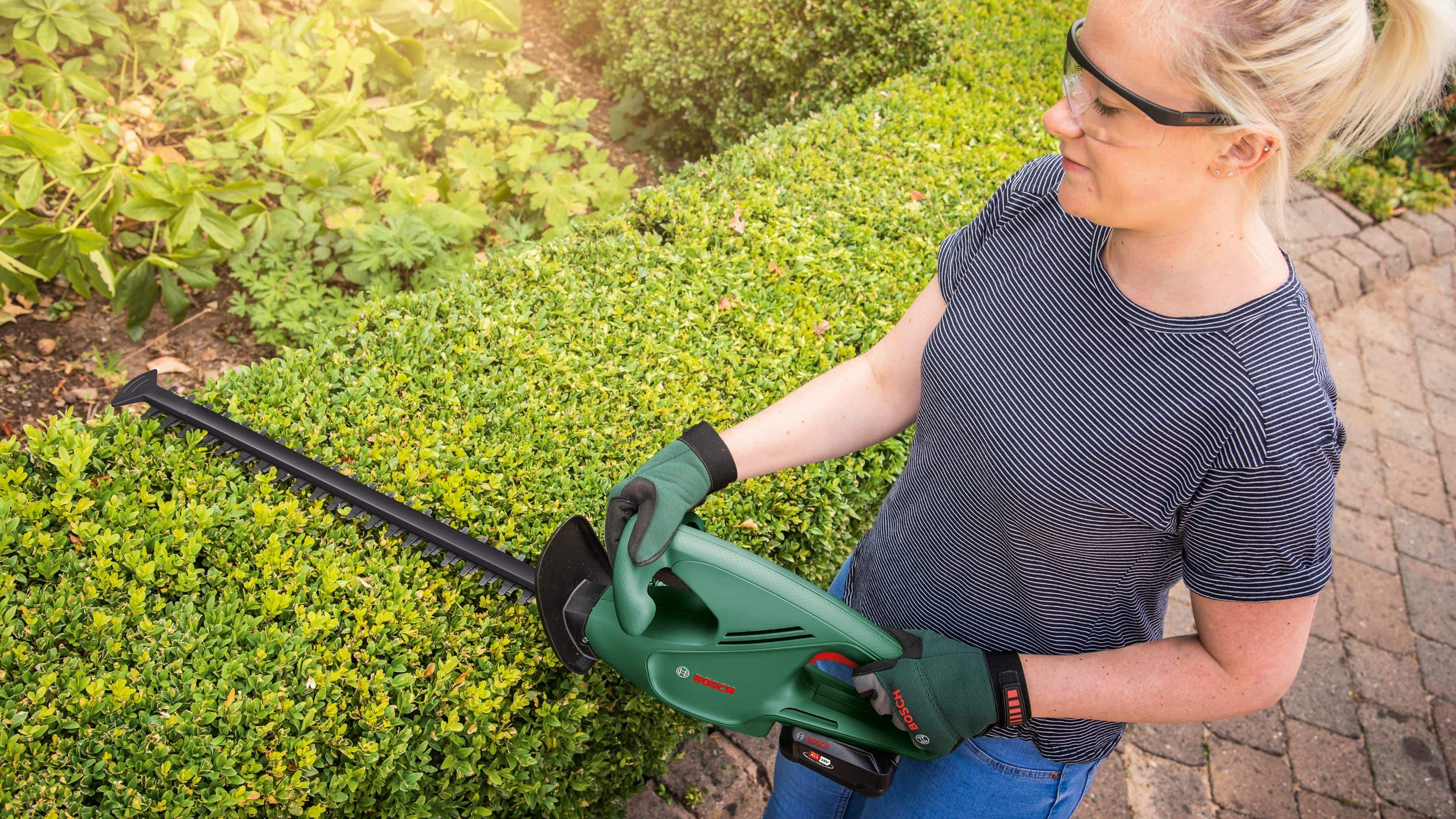 Kobieta używa zielonych nożyc do żywopłotu na zielonym żywopłocie. Nosi okulary ochronne i rękawice. W tle rośliny.