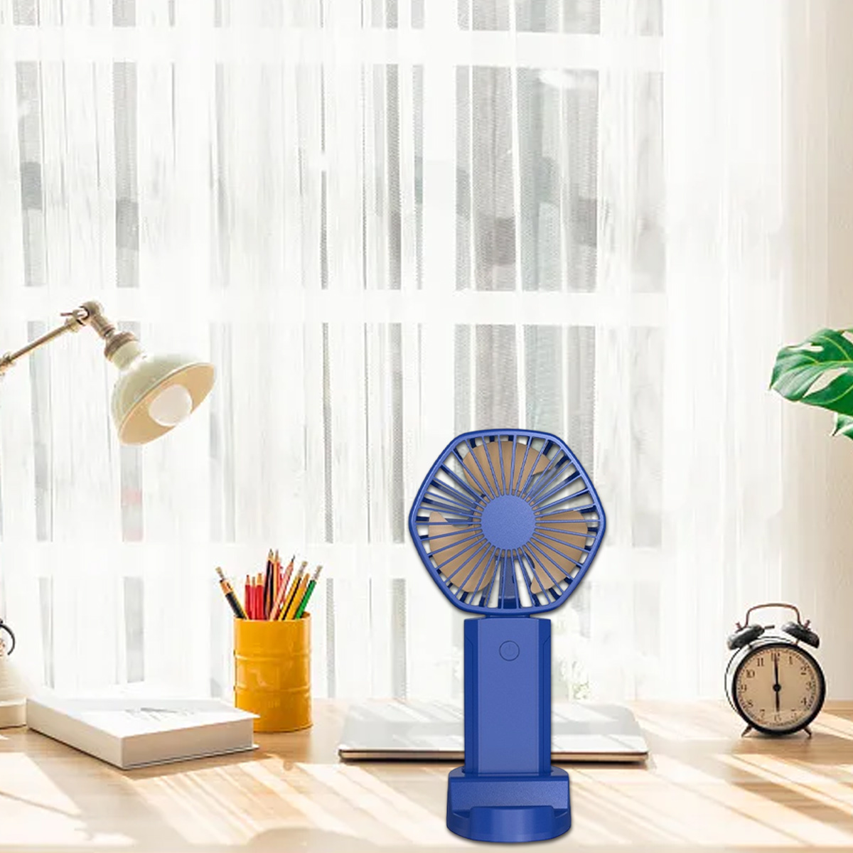 Ein blauer tragbarer Ventilator auf einem Schreibtisch mit Bleistiften, einer Lampe und einem Wecker.
