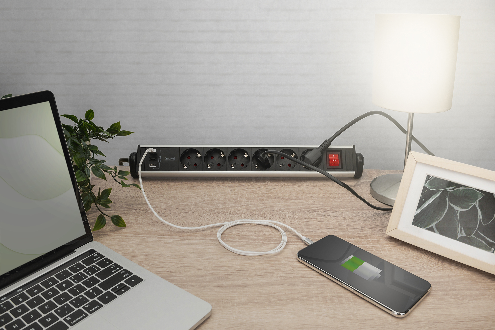 Laptop, ładowanie telefonu, lampa i ramka na zdjęcia na drewnianym biurku. Listwa zasilająca jest w centrum uwagi.
