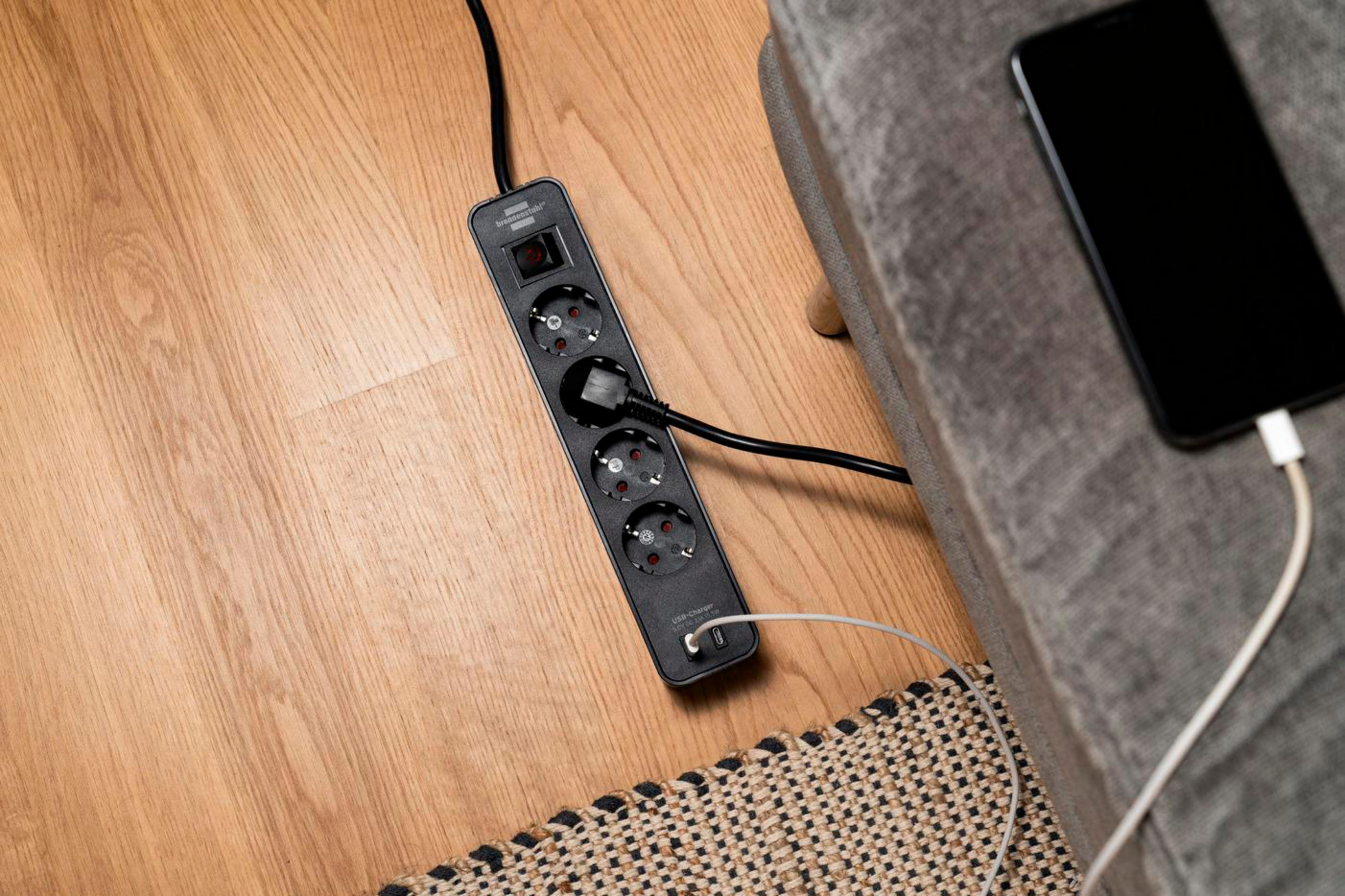 Czarna listwa zasilająca z czterema gniazdami, przełącznikiem i ładowarką USB, umieszczona na drewnianej podłodze.