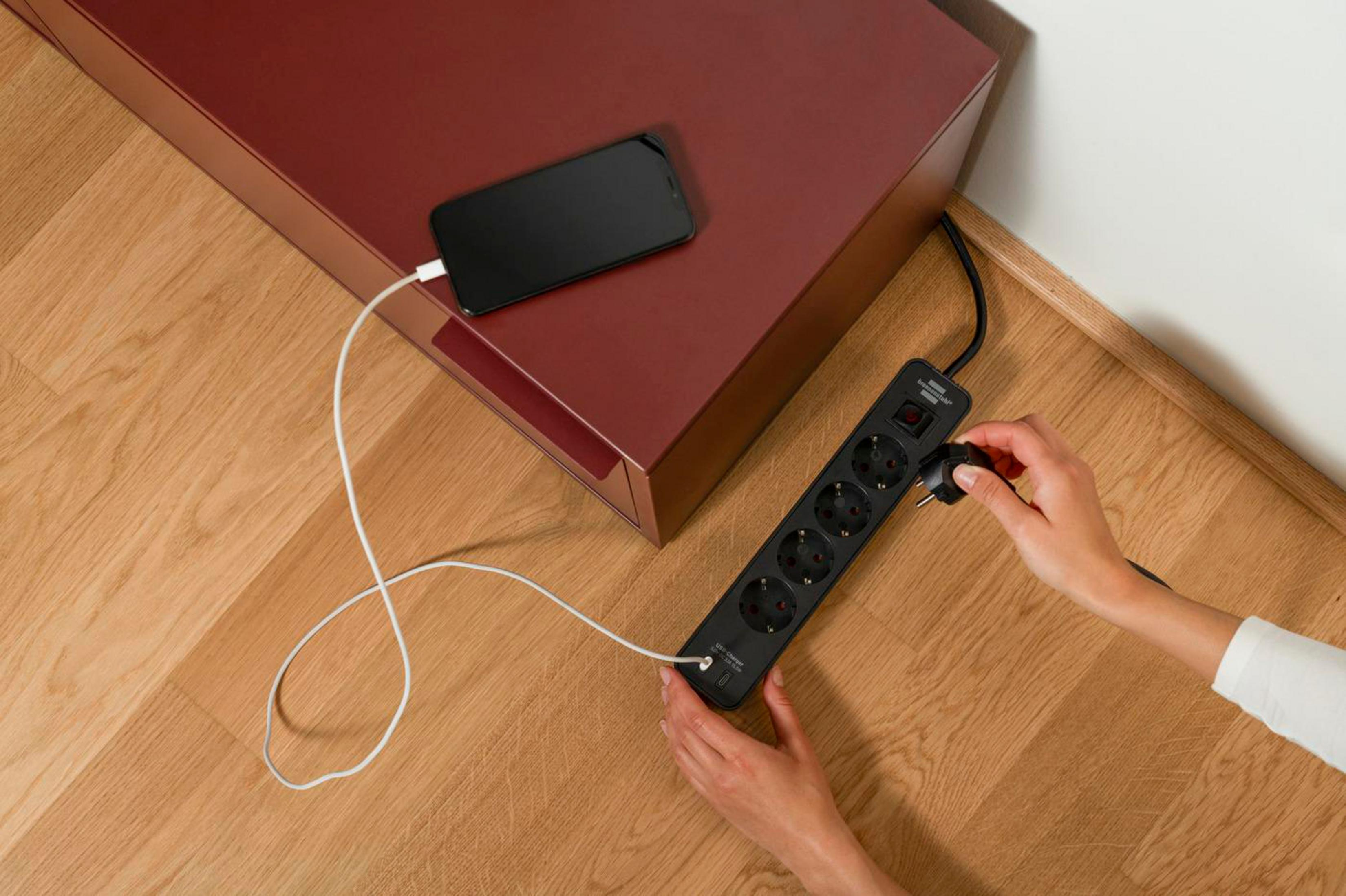 Telefon ładujący się na czerwonej szafce, podłączony do listwy zasilającej na drewnianej podłodze.