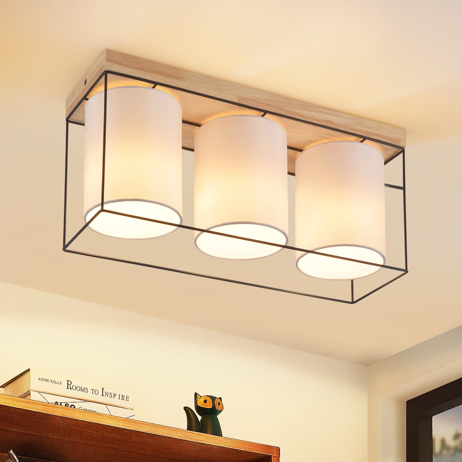 Un apparecchio di illuminazione a soffitto con tre paralumi cilindrici bianchi, cornice nera e piano in legno.