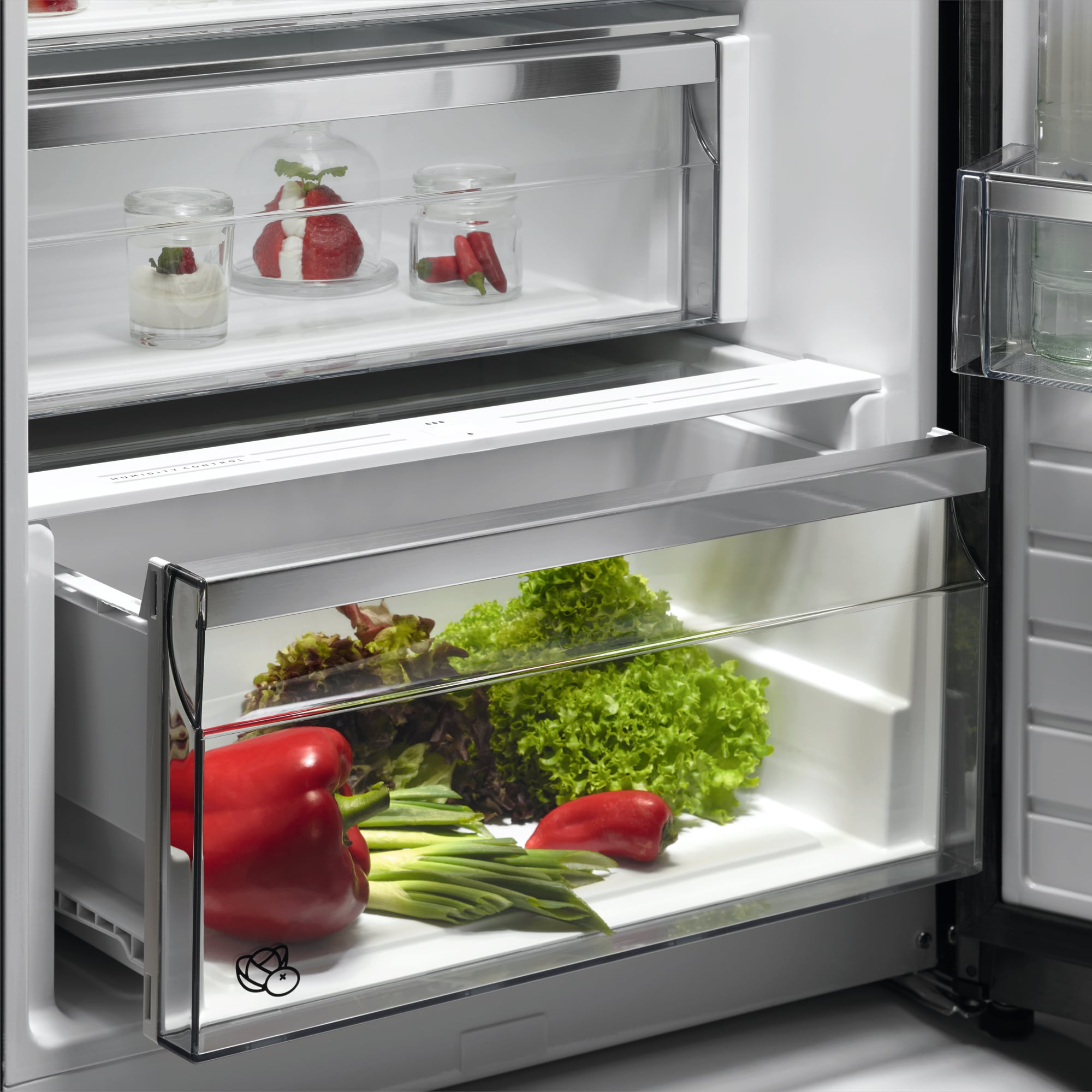Offener Kühlschrank mit Schubladen voller Gemüse und Gläsern mit Obst. Rote Paprika, Salat und Erdbeeren sind sichtbar.