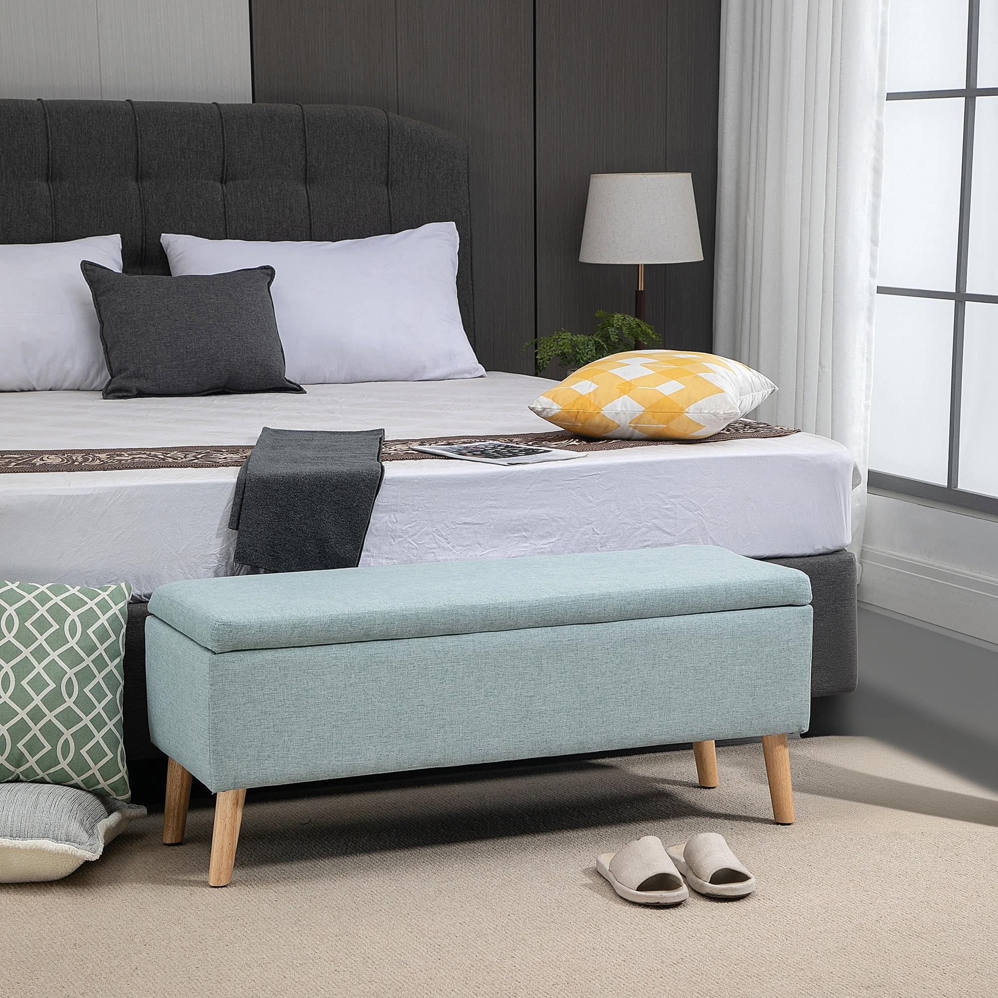 Eine blaue Aufbewahrungsbank mit Holzbeinen in einem Schlafzimmer. Kissen und eine Lampe befinden sich auf dem Bett.