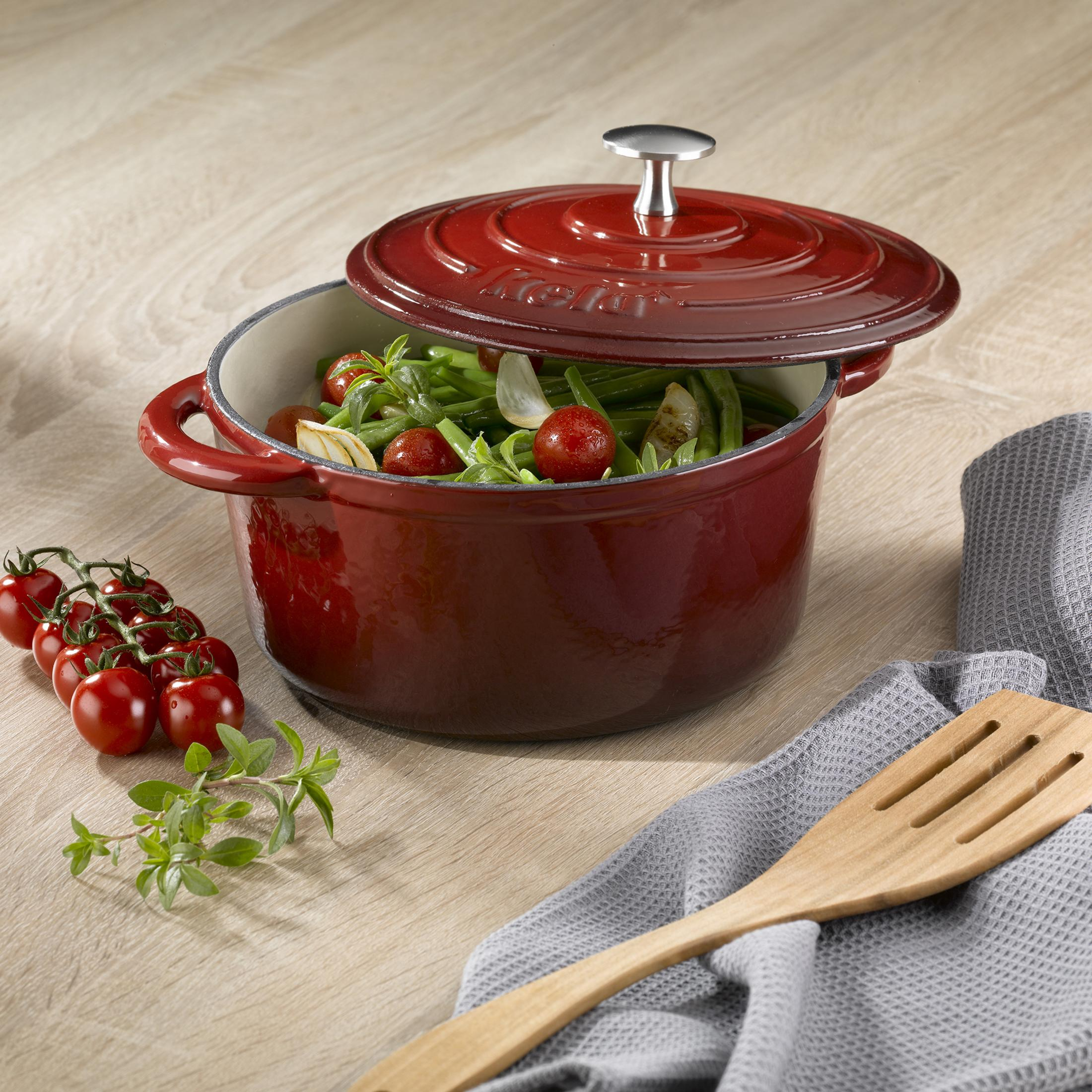 Roter Kela-Topf mit offenem Deckel, gefüllt mit Gemüse und Tomaten. Holzlöffel und graues Tuch.