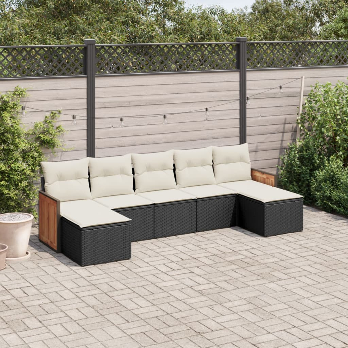 Divano componibile in rattan nero con cuscini bianchi. Sfondo: pietre del patio, recinzione e piante.