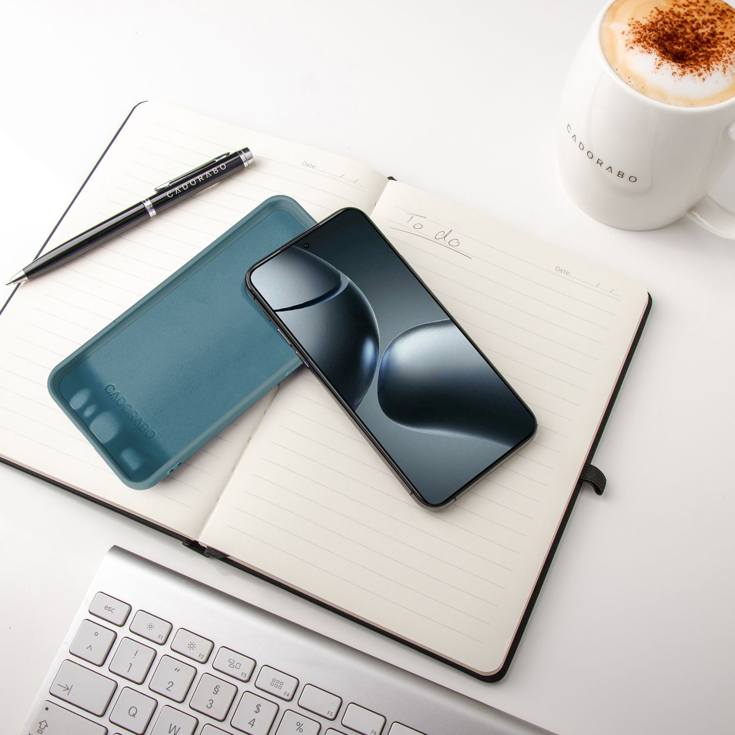Bild: Türkisfarbene Handyhülle, Telefon, Stift und Kaffeetasse auf einem Notizbuch mit Tastatur.