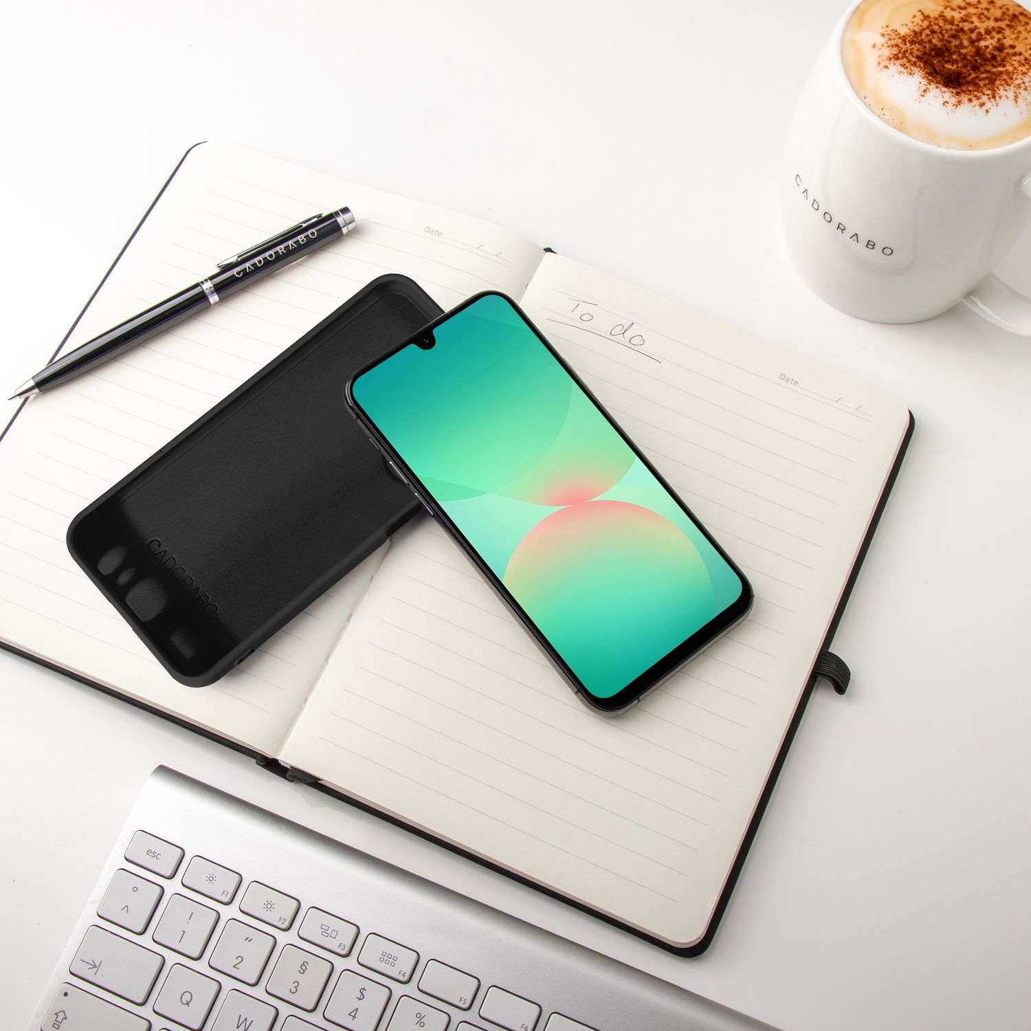 Ein schwarzes Telefon und eine Hülle auf einem Notizbuch. Ein Stift und eine Kaffeetasse stehen daneben, auf einem weißen Tisch.