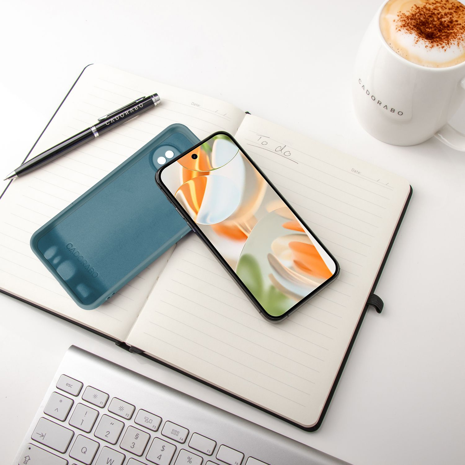 Telefon in türkisfarbener Hülle, offenes Notizbuch, Stift und Kaffeetasse auf weißer Oberfläche. Tastatur im Hintergrund.
