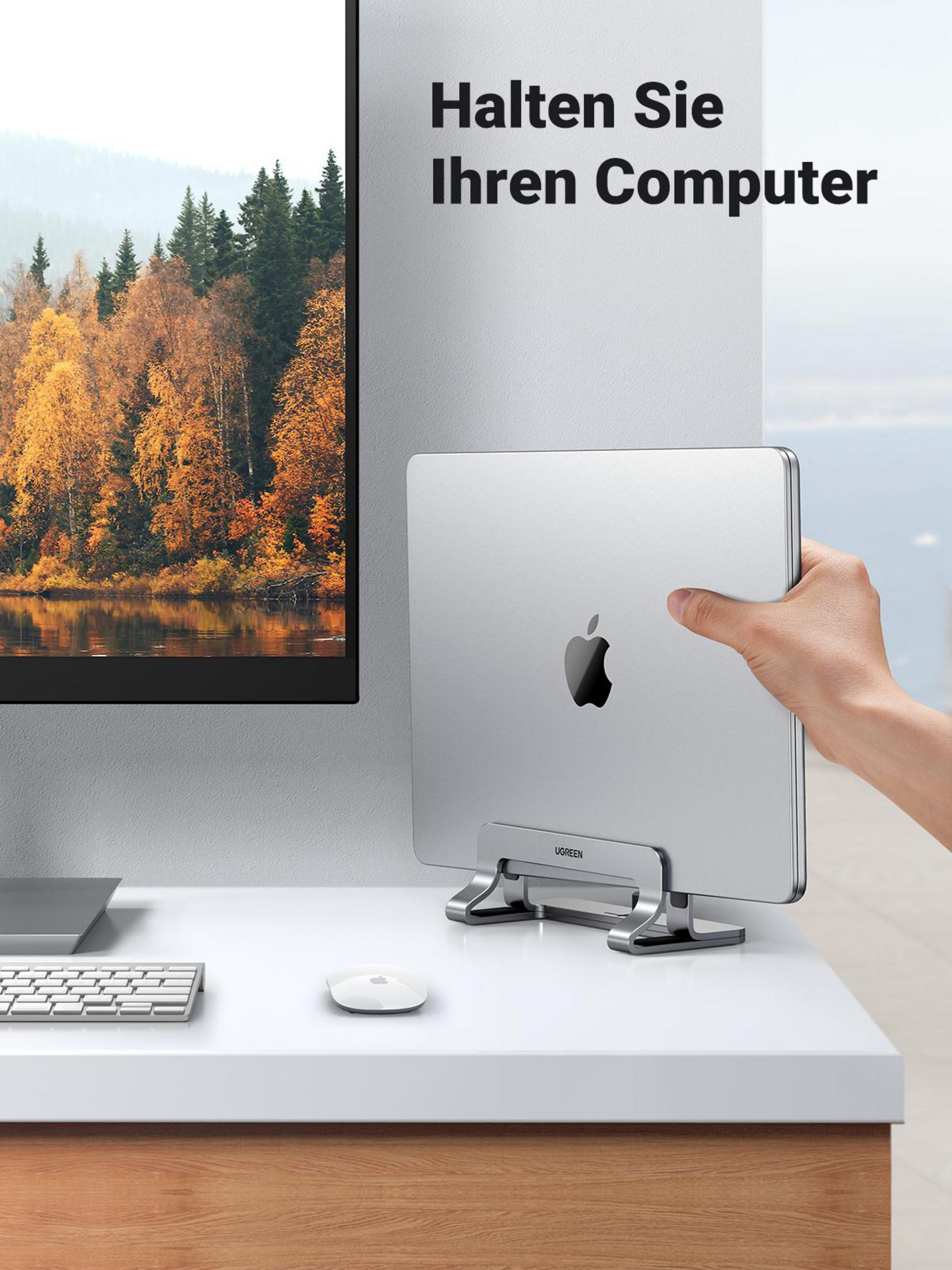 Person hält ein MacBook in einem silbernen Laptopständer neben einem Monitor.