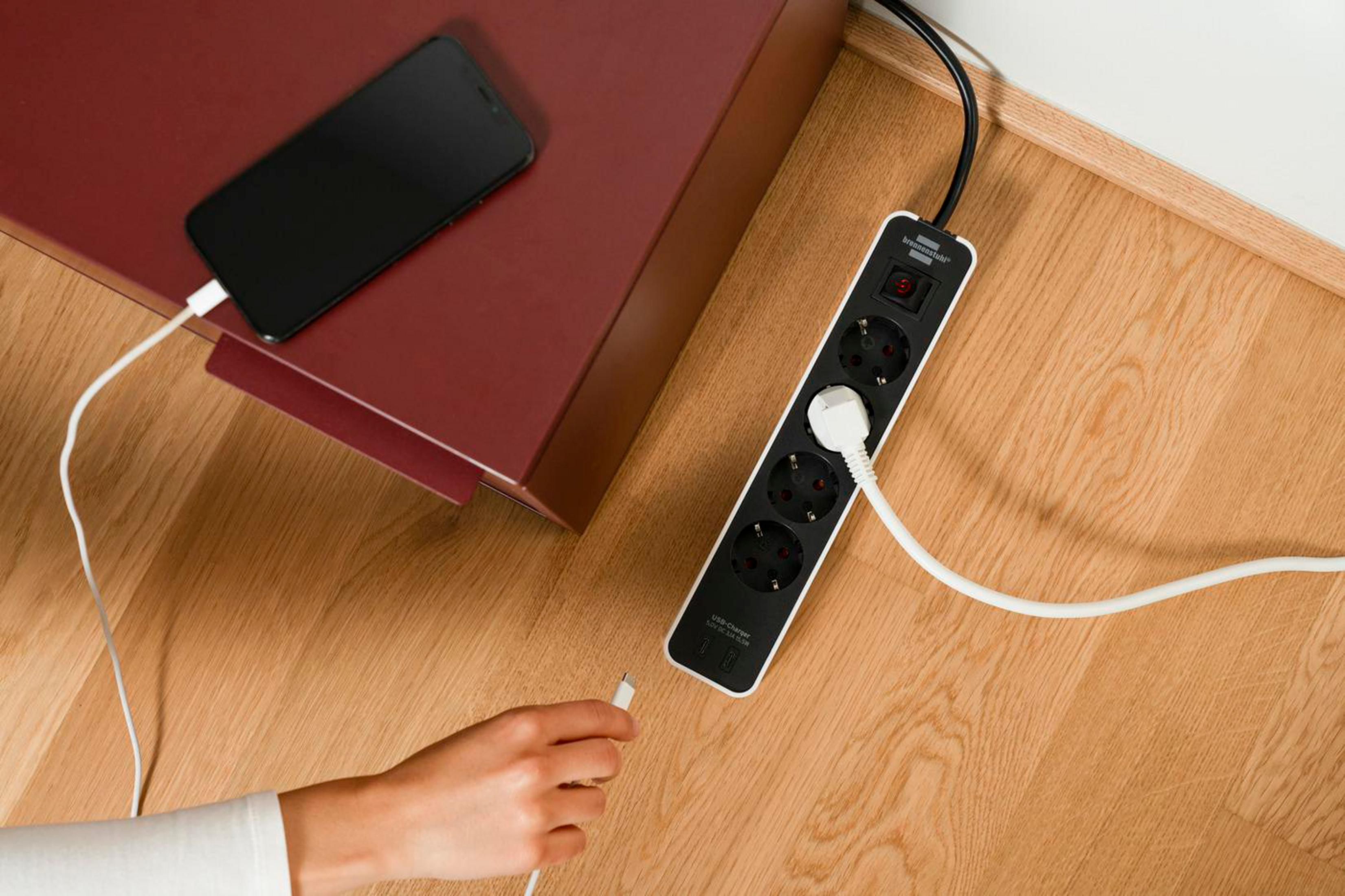 Ręka trzymająca kabel ładujący obok czarnego telefonu na listwie zasilającej, na drewnianej podłodze.