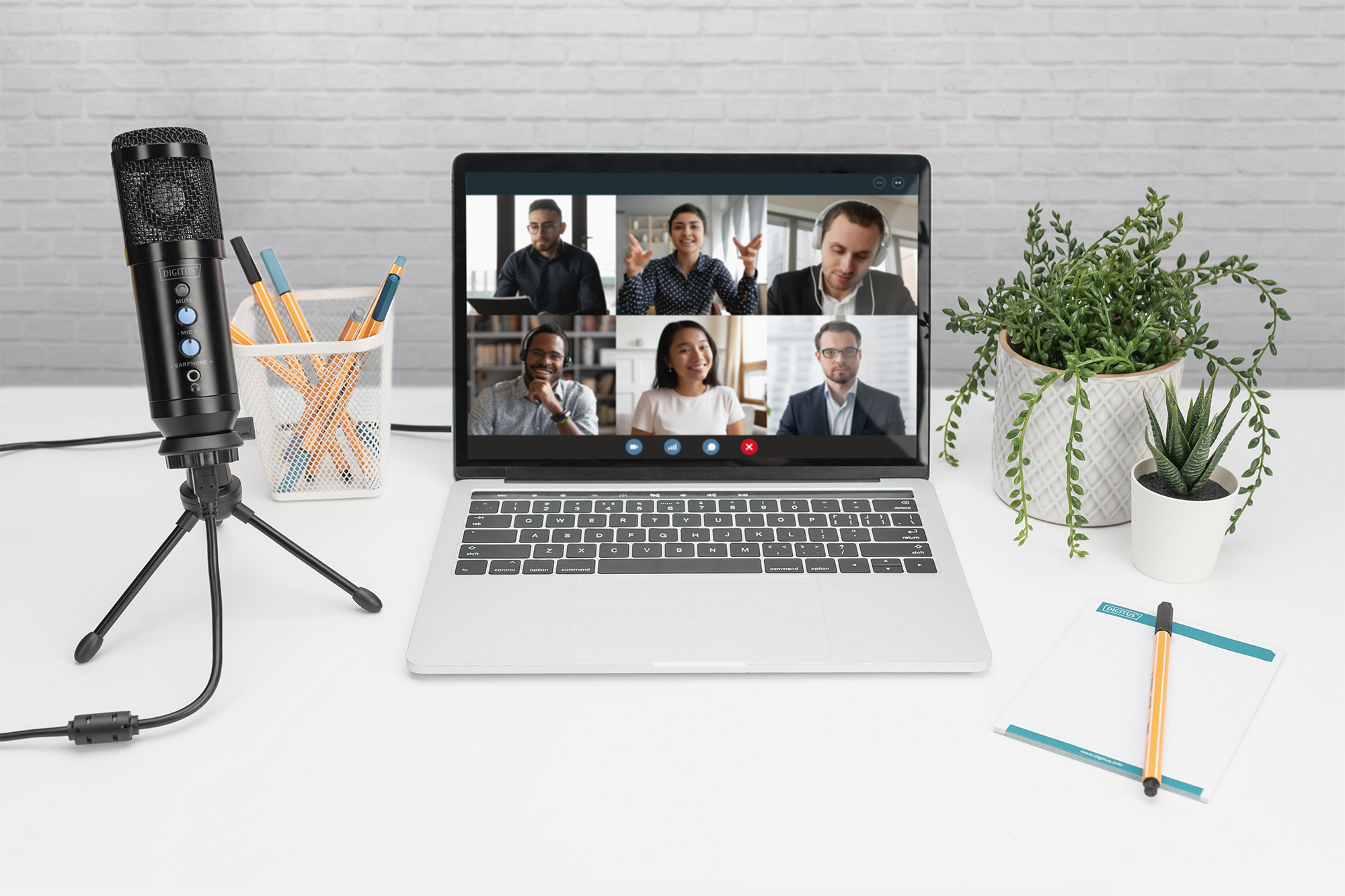Laptop che mostra una videochiamata con 6 persone. Visibili un microfono e forniture per ufficio.