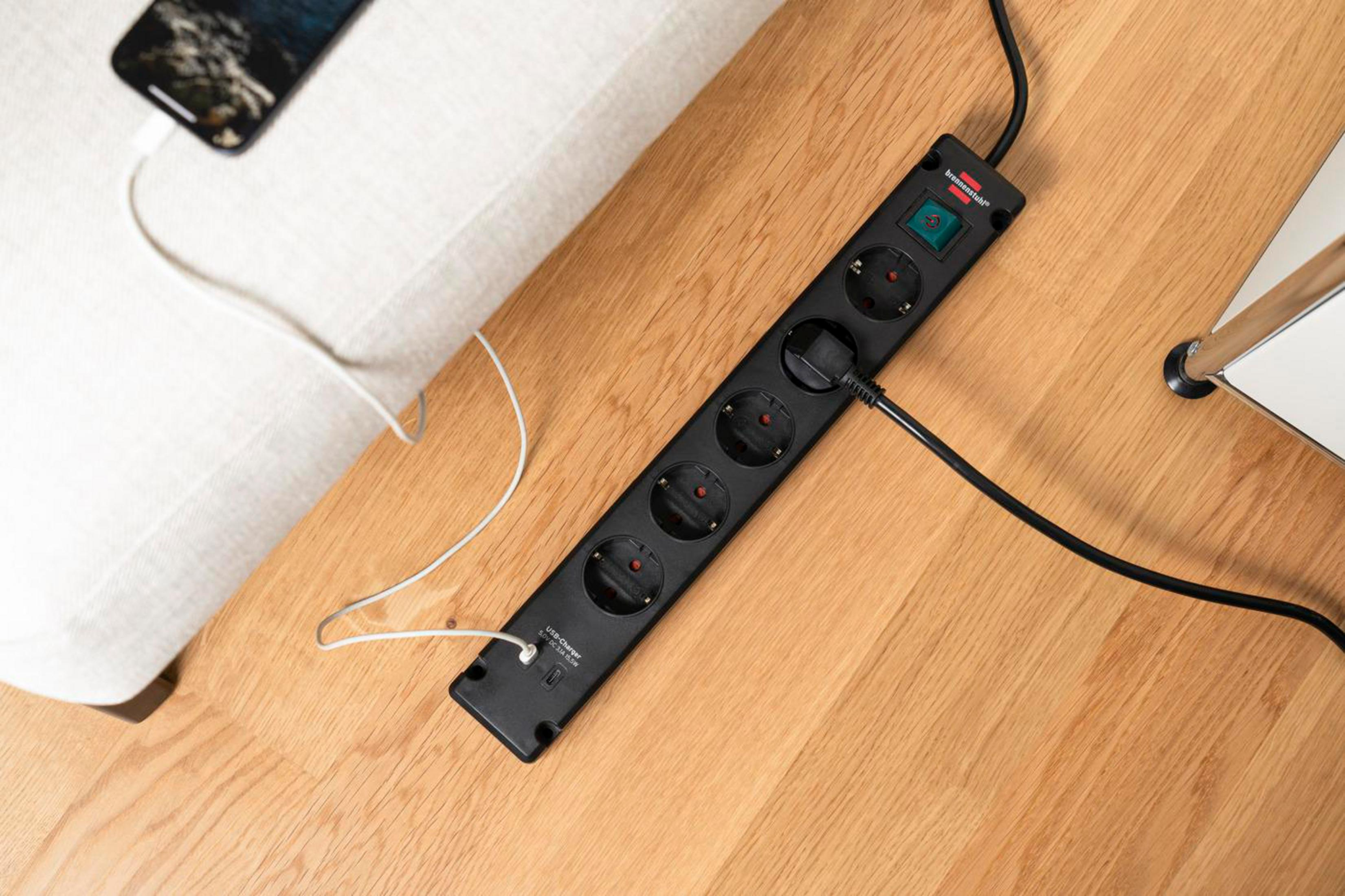 Czarny przedłużacz na drewnianej podłodze, z telefonem w trakcie ładowania. Listwa ma 5 gniazd.
