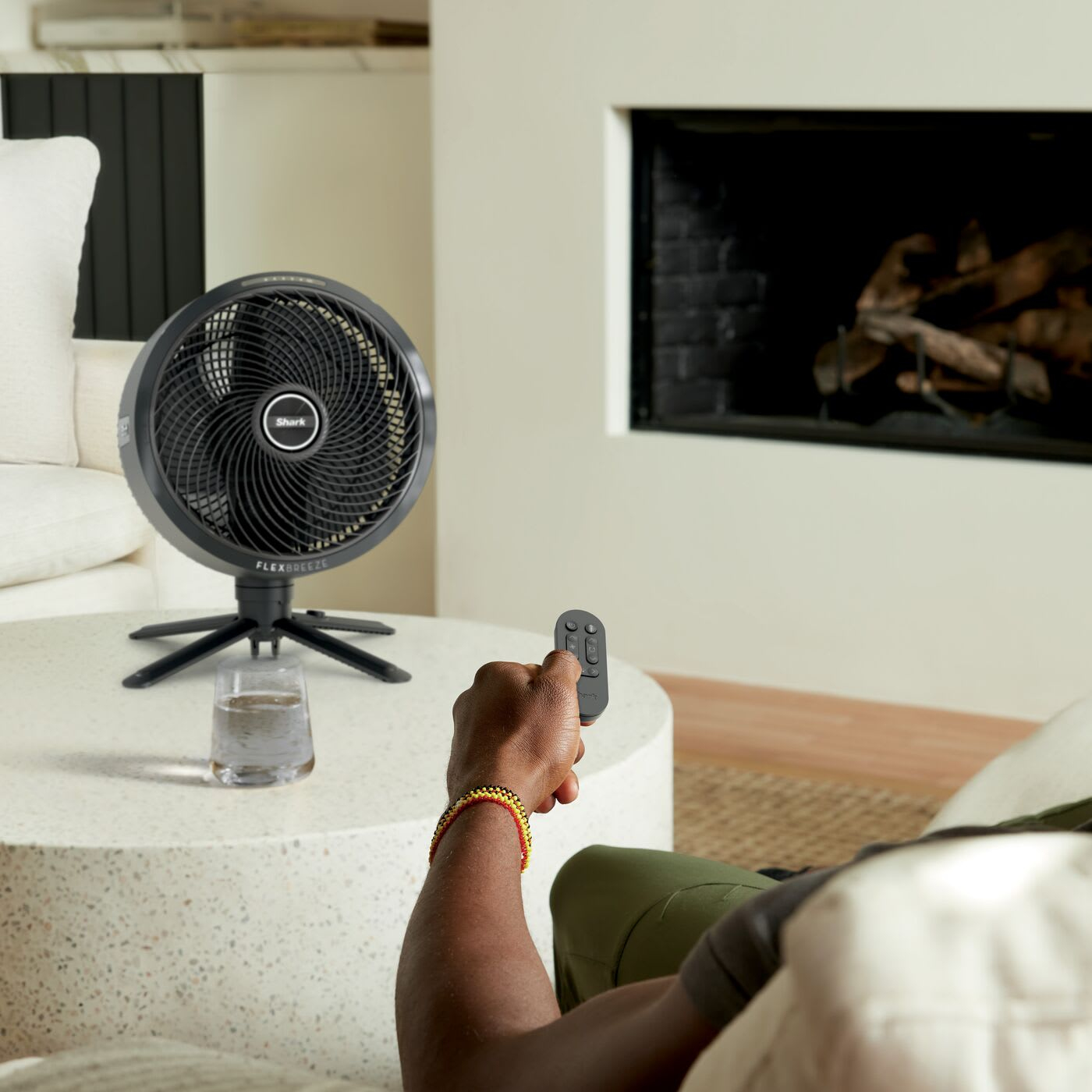 Osoba z pilotem. Wentylator na stole, szklanka wody, kominek w tle.