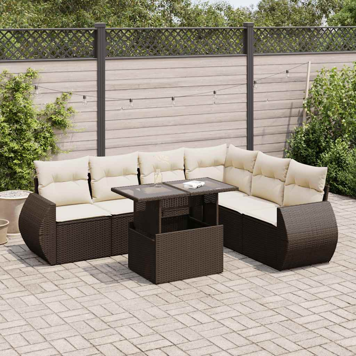 Set patio esterno: divano in vimini marrone, tavolo e cuscini. Recinzione in legno e sfondo verde.
