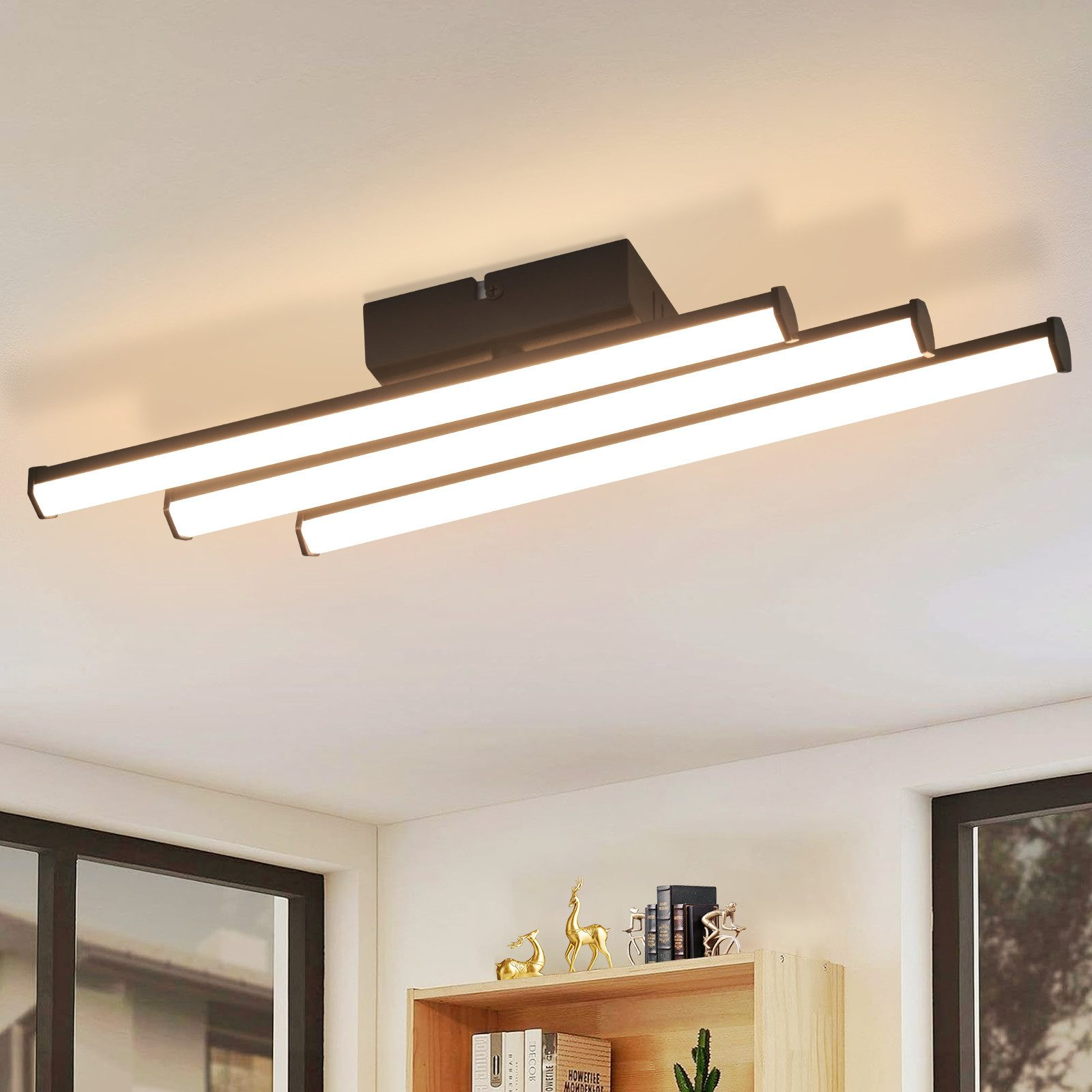 Moderne schwarze Deckenleuchte. Vier Lichtbalken auf schwarzem Sockel. Weiße Decke. Die Lichter sind an.