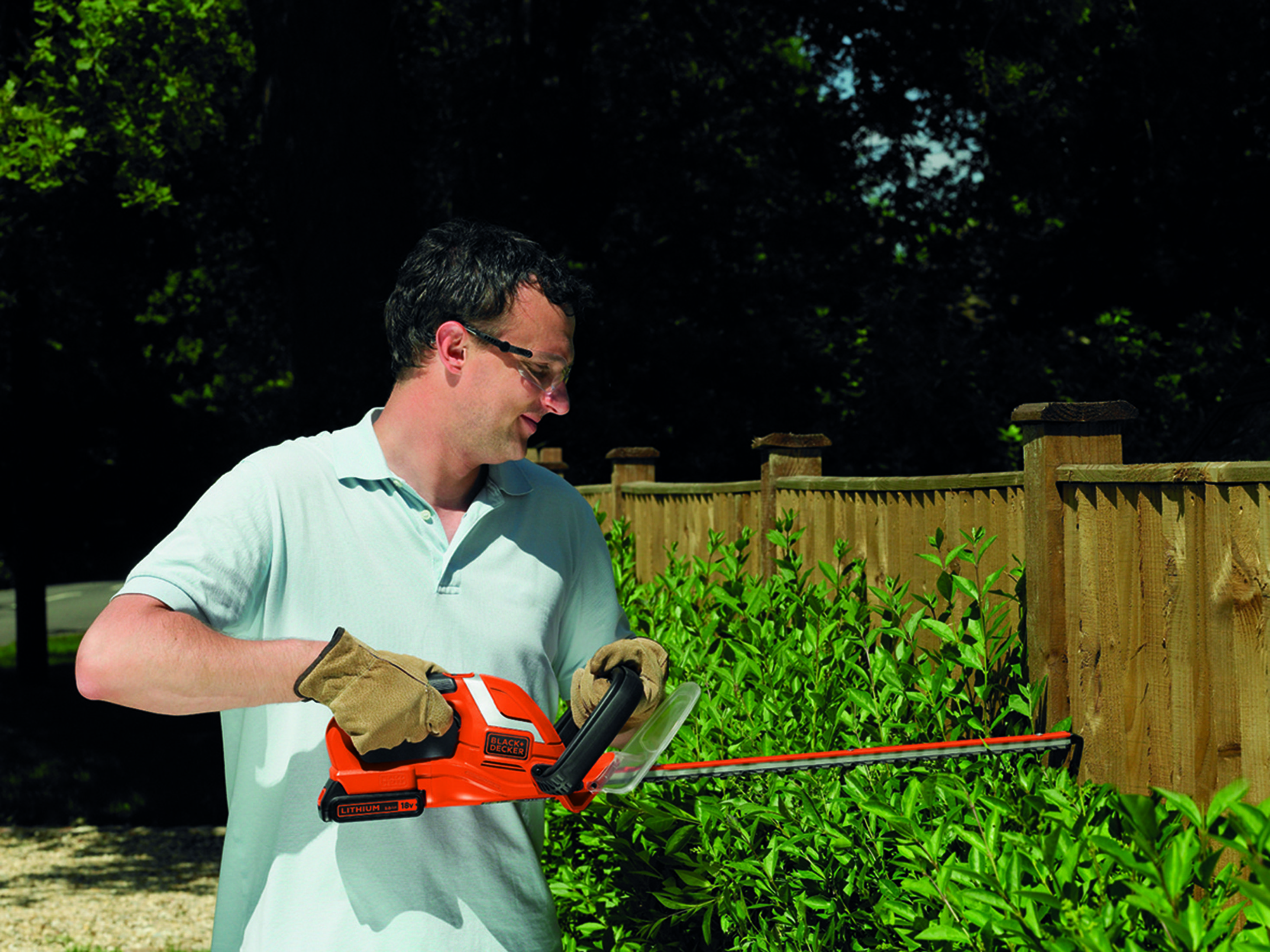 Mężczyzna w jasnoniebieskiej koszuli przycina krzewy pomarańczowo-czarnymi nożycami do żywopłotu.