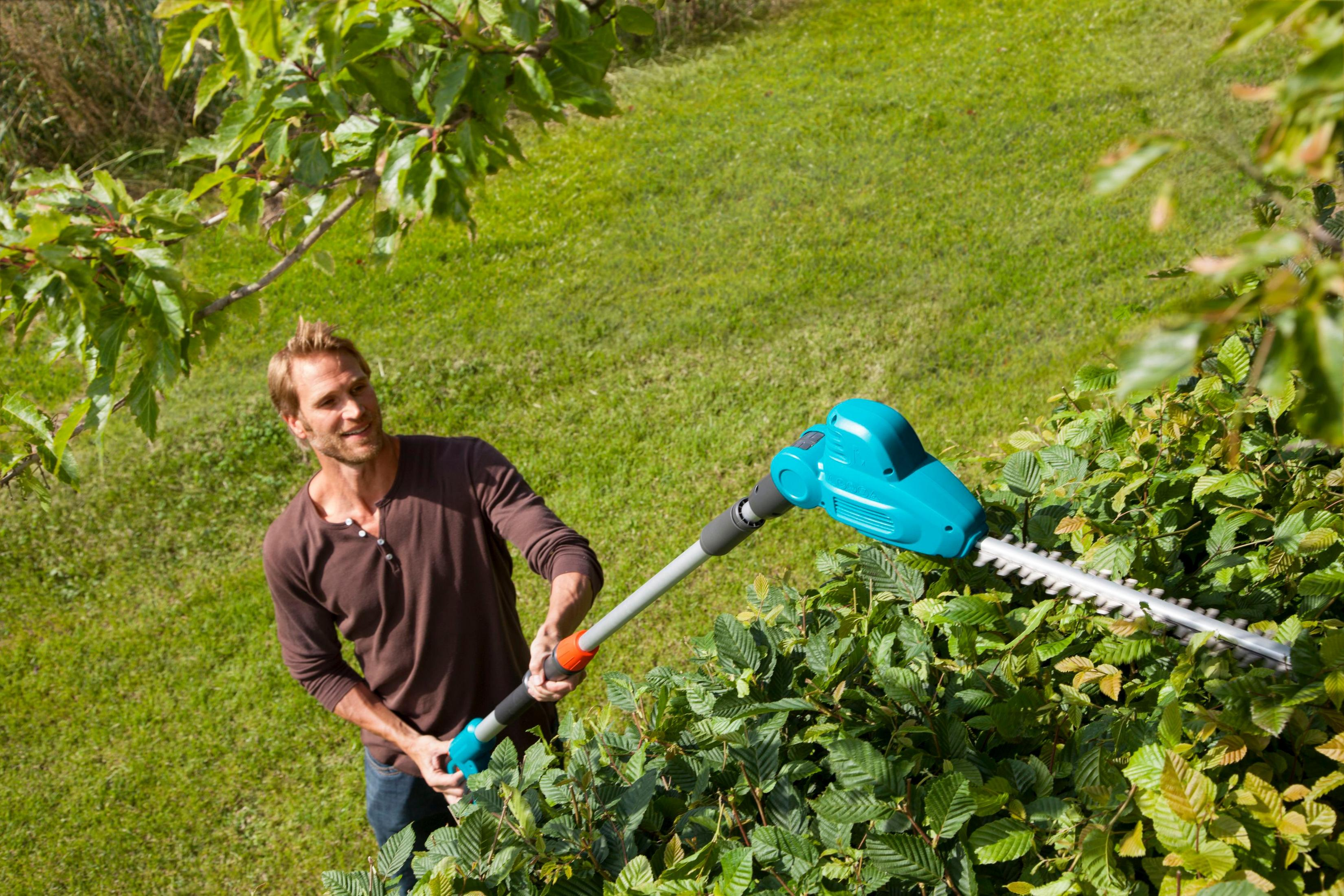 Mann schneidet Hecke mit langstieligem elektrischen Trimmer. Grüne Büsche und Bäume im Hintergrund.
