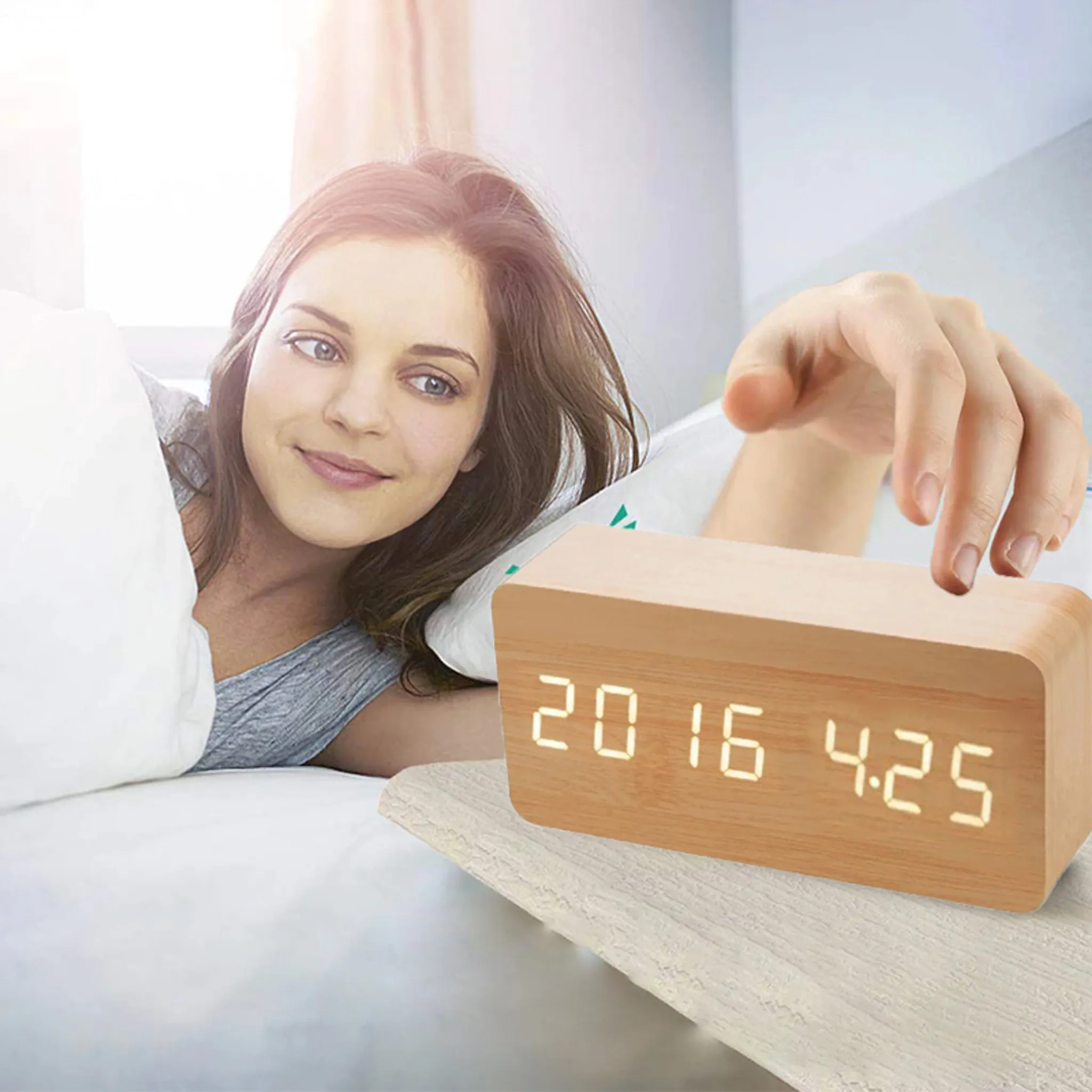 Eine Frau im Bett greift nach einer digitalen Holzuhr. Die Uhr zeigt ein Datum.