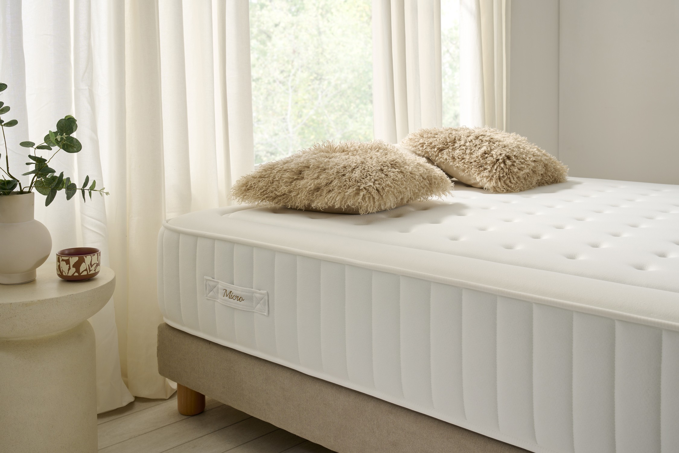 Colchón blanco con almohadas mullidas, ventana al fondo, tonos neutros.