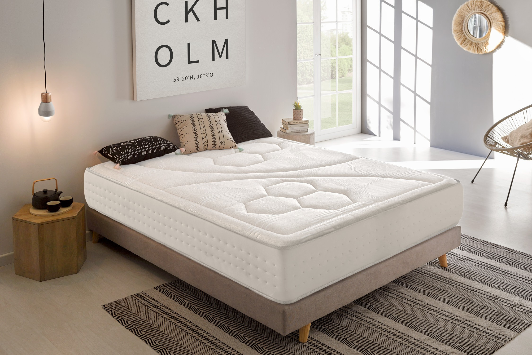Una cama con colchón blanco, almohadas y una mesita de noche de madera. Tonos neutros.