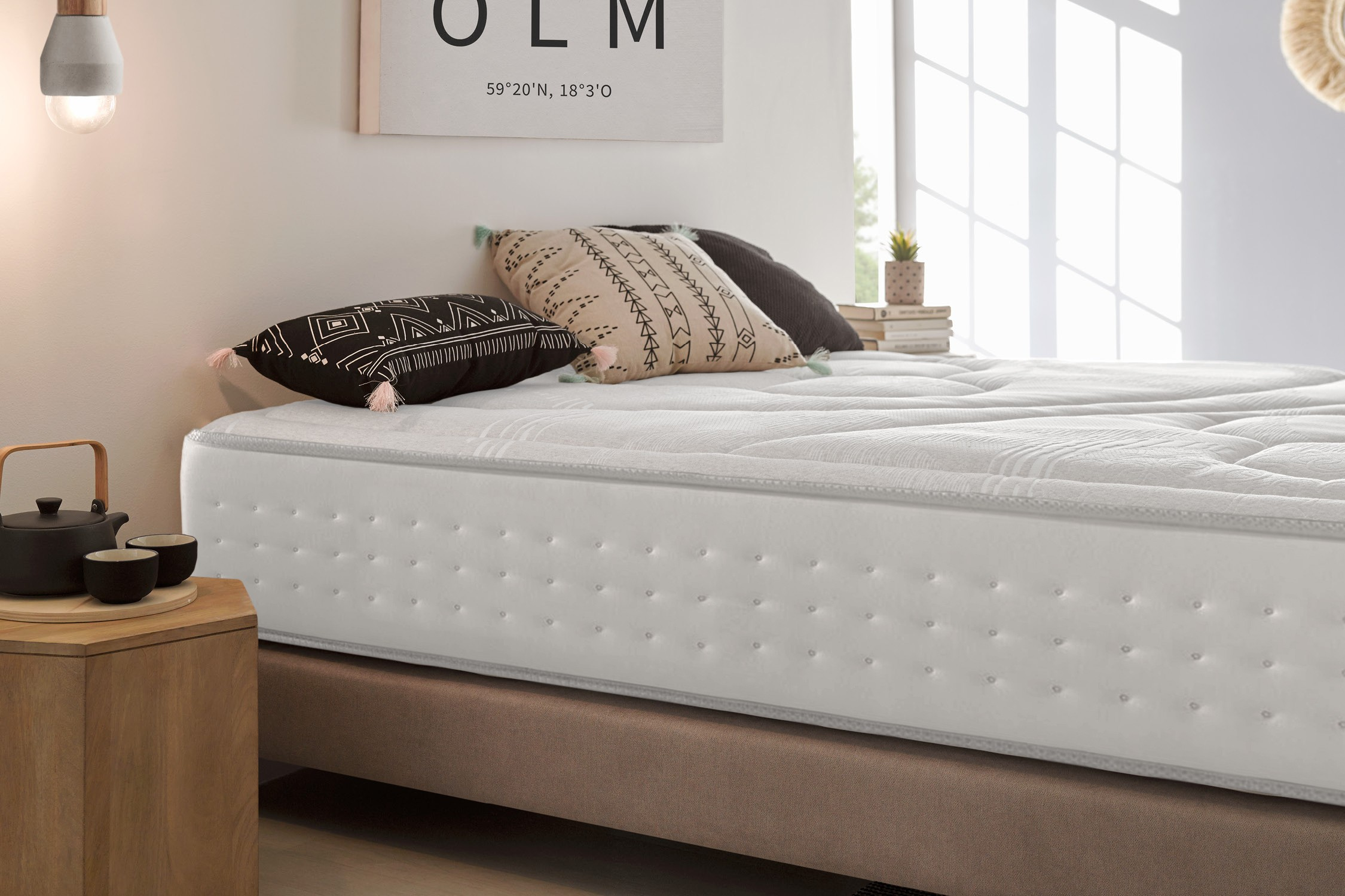 Una cama con colchón blanco, almohadas y una mesita de noche de madera. Tonos neutros.