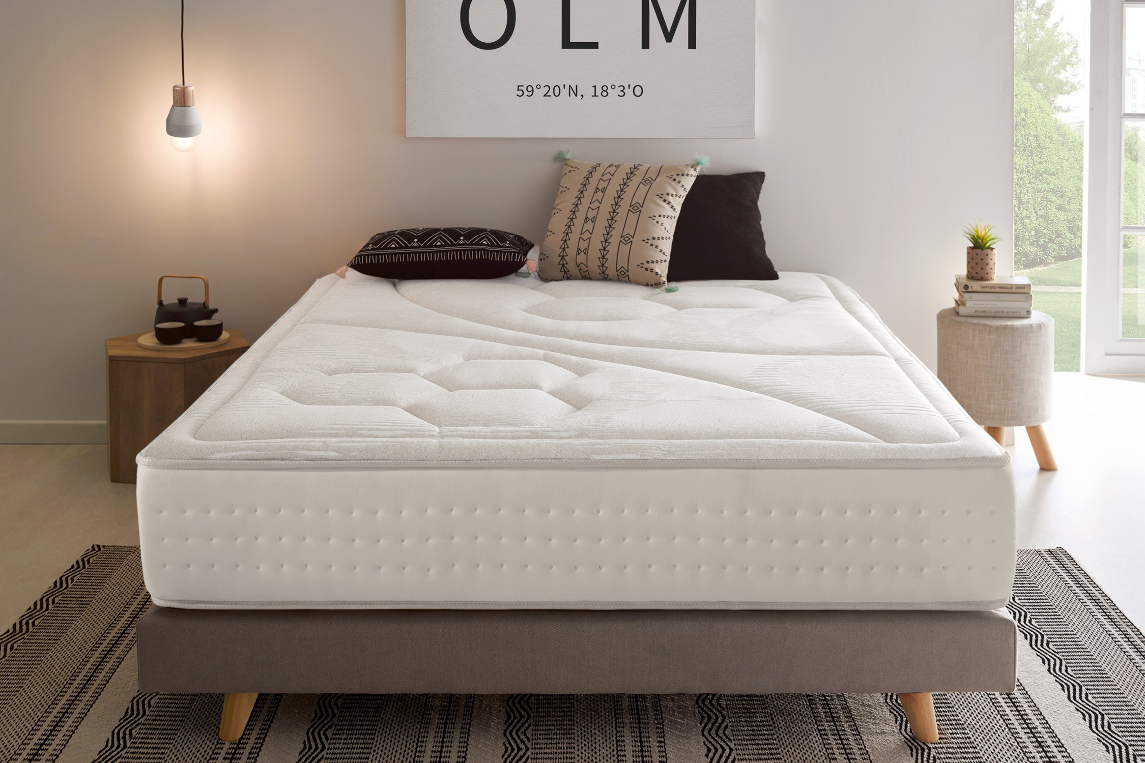 Una cama con colchón blanco, almohadas y una mesita de noche de madera. Tonos neutros.