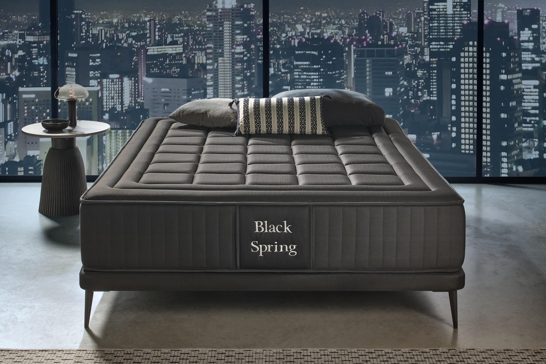 Cama negra con almohadas y vista nocturna de una ciudad desde una ventana grande.
