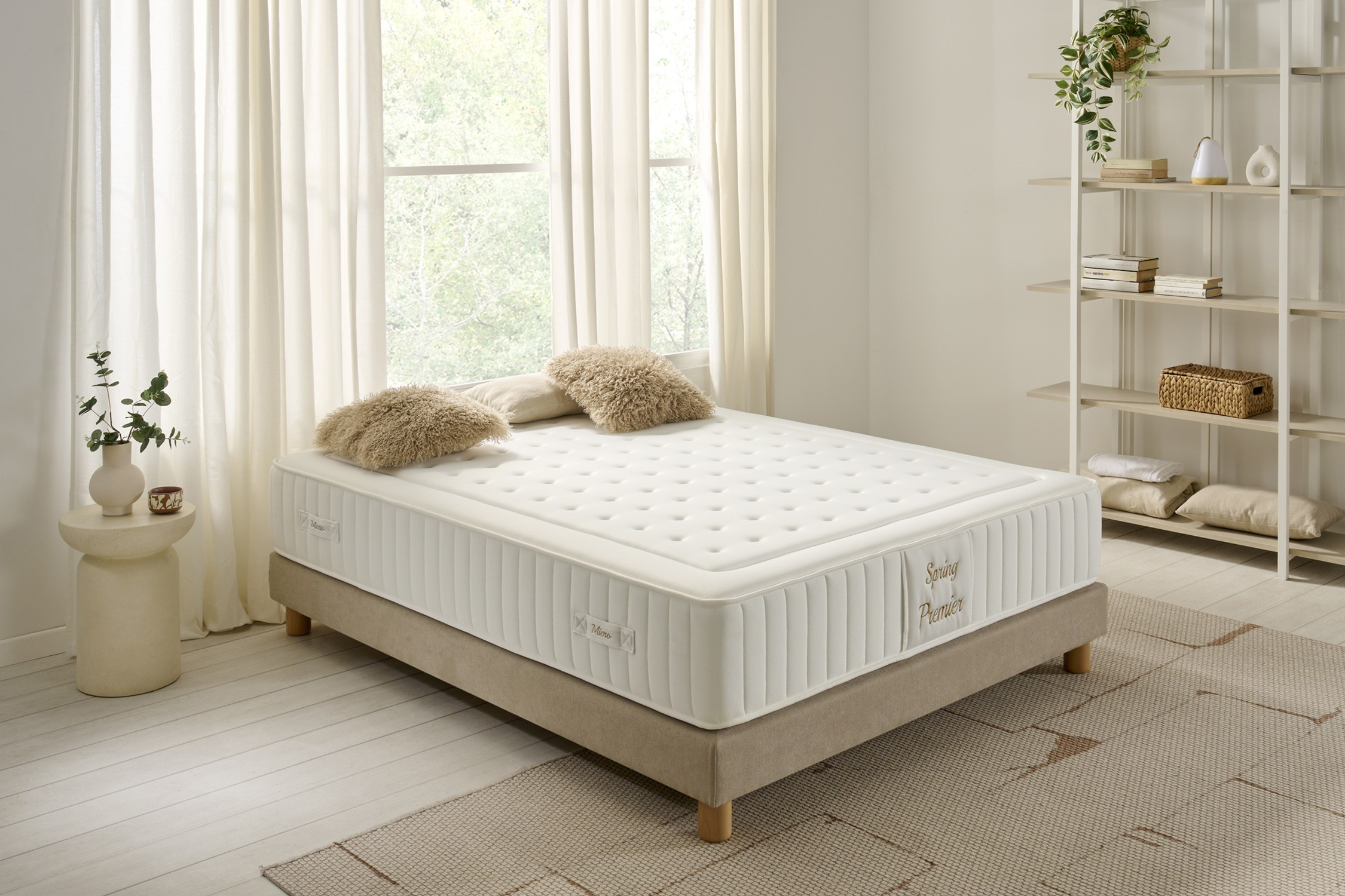 Cama con colchón, almohadas y base beige. Ventana y estanterías al fondo.
