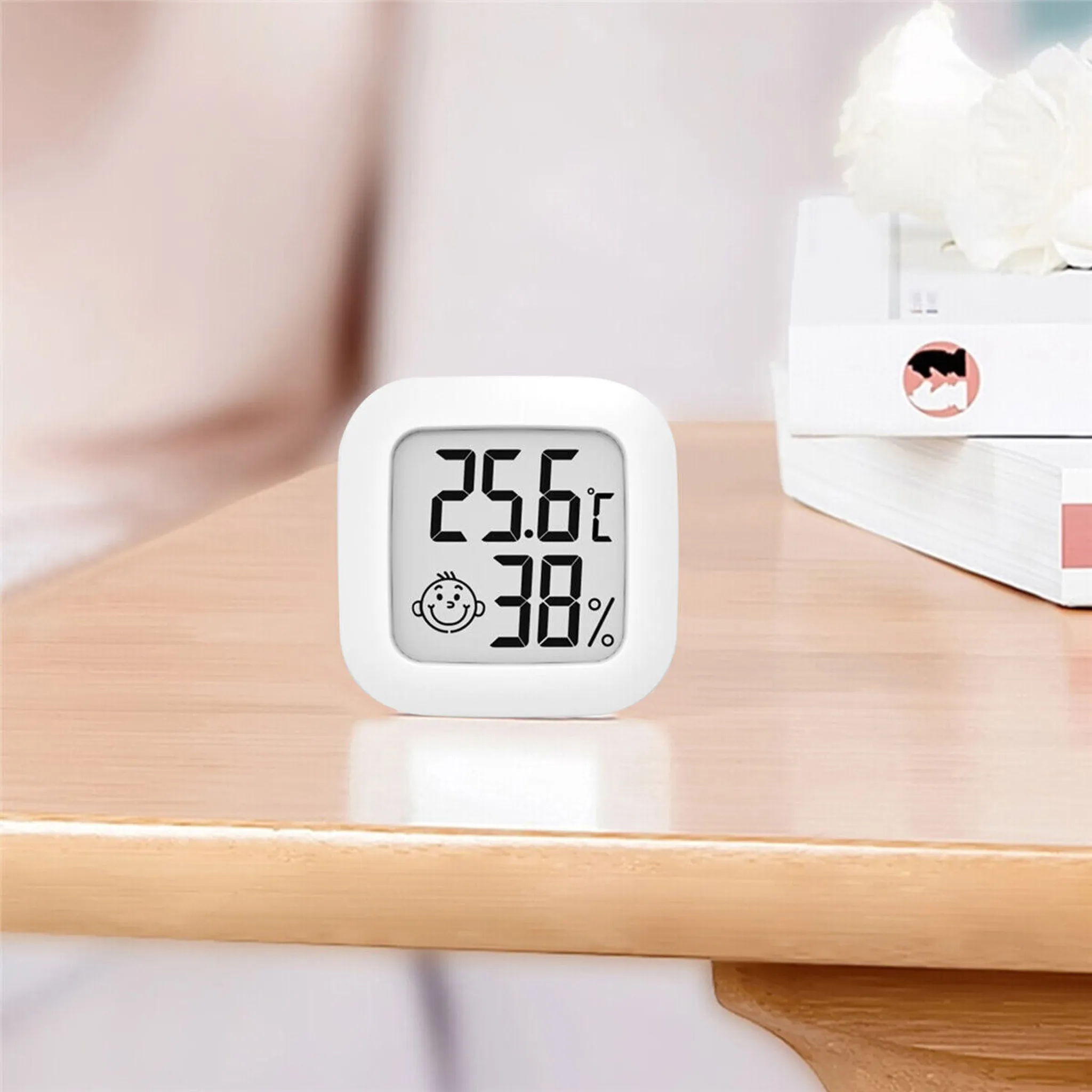 Ein weißes digitales Thermometer auf einem Holztisch, das 25,6 °C und 38 % anzeigt. Daneben Bücher und Blumen.