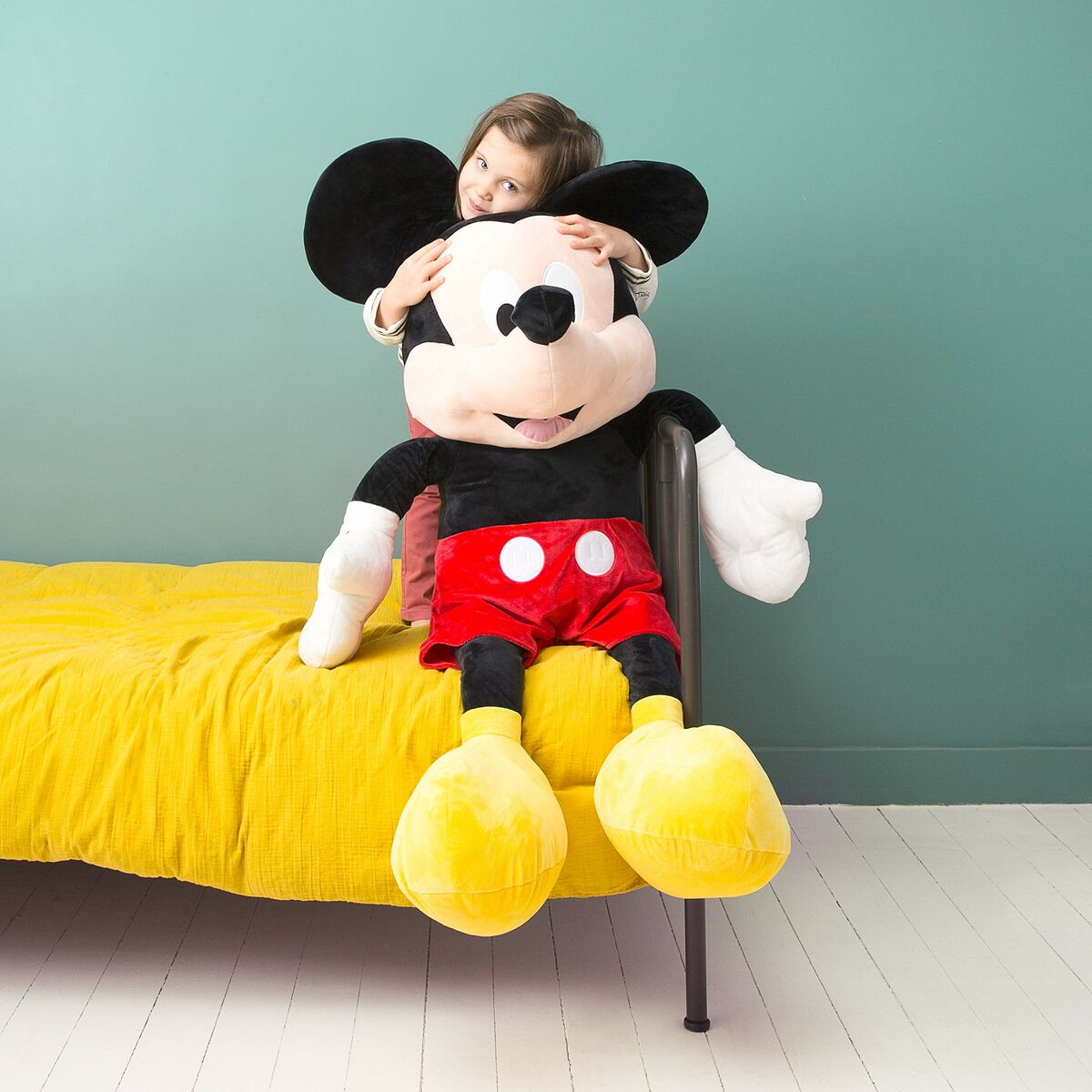 Niño abrazando peluche de Mickey Mouse. Colcha amarilla, fondo de pared verde.