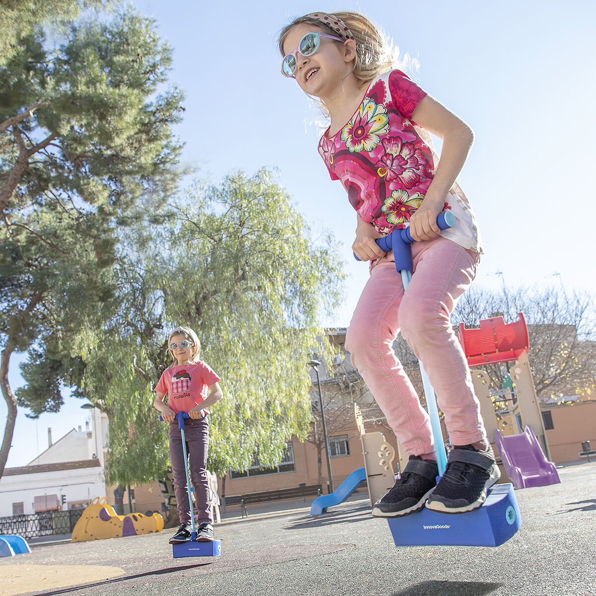 Dos niñas saltan en pogo sticks en un parque, una en el aire. Los árboles y el equipo de juego están al fondo.