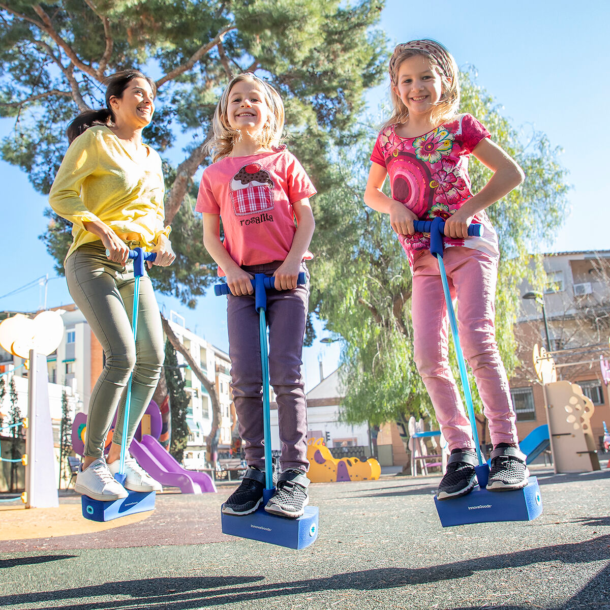 Tres personas saltan en pogo sticks en un parque. Una mujer y dos niñas. Edificios y un parque infantil en el fondo.