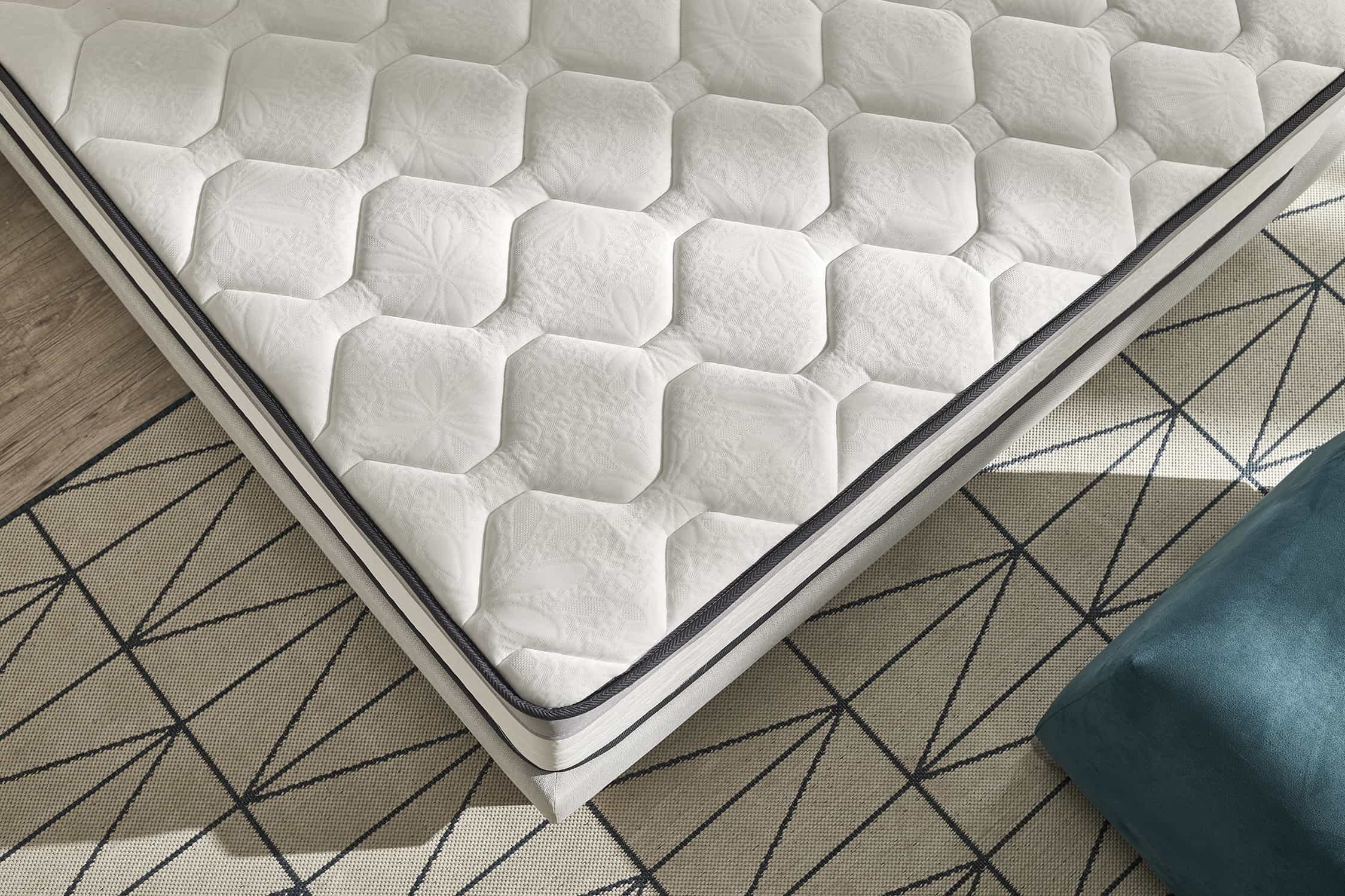 Colchón blanco con patrón de diamantes, sobre una alfombra estampada. Cojín azul parcialmente visible.