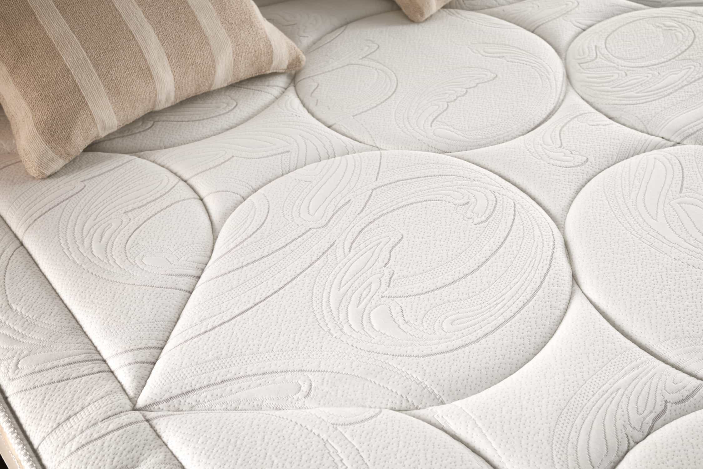 Primer plano de un colchón blanco con diseño acolchado y dos almohadas a rayas en el fondo.