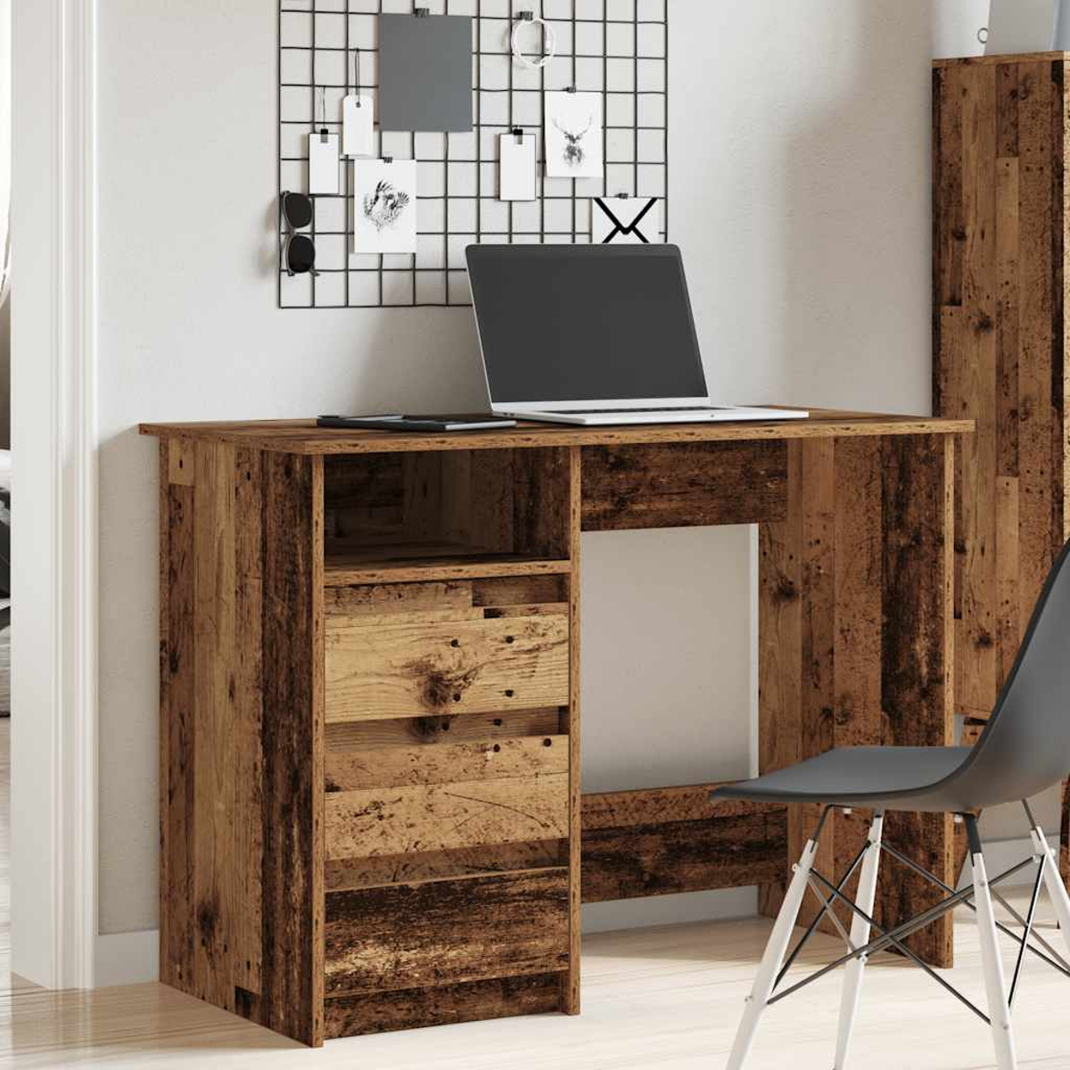Holzschreibtisch an einer Wand, mit Schubladen und Laptop. Schwarzes Gitter mit Notizen sichtbar.