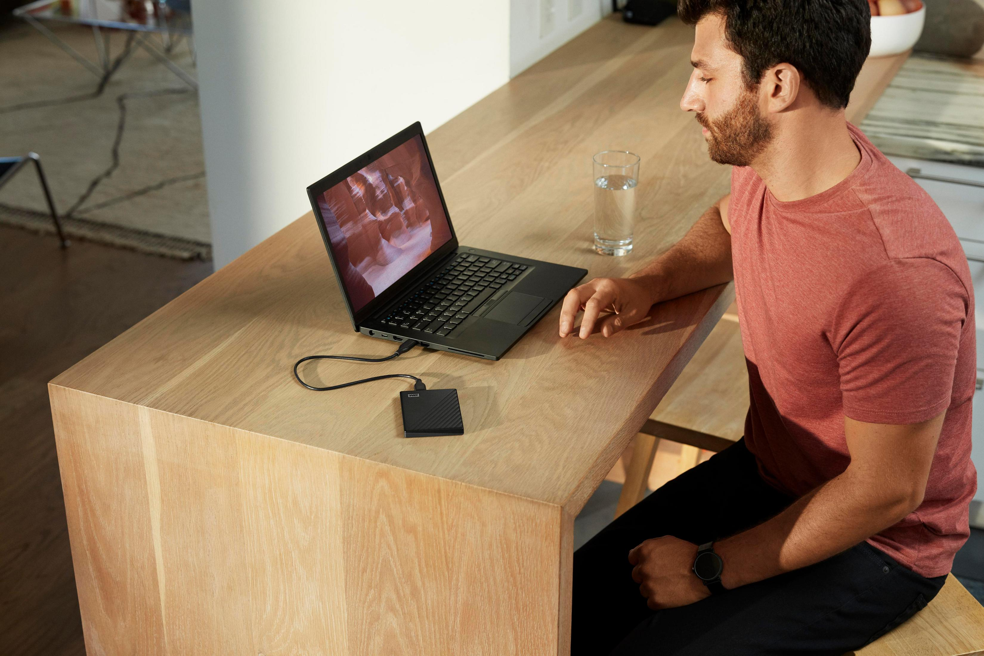 Mężczyzna używający laptopa z podłączonym dyskiem twardym, siedzący przy drewnianym stole z szklanką wody.