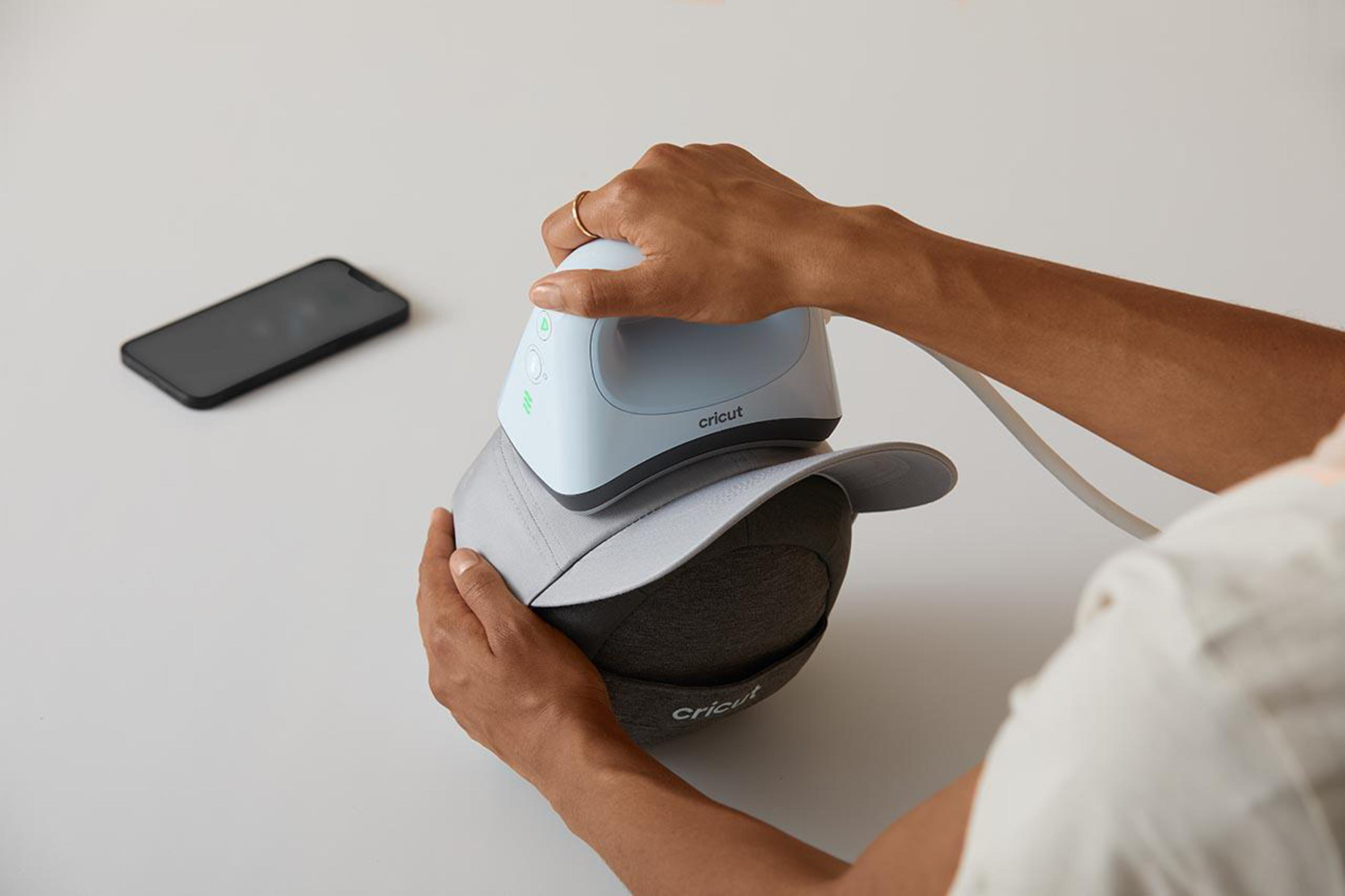 Mani che usano una pressa a caldo Cricut su un cappellino da baseball grigio. Un telefono è vicino.